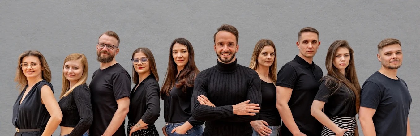 Zespół agencji marketingowej w czarnych strojach na tle szarej ściany. Mężczyzna w golfie na pierwszym planie, uśmiechnięty, z założonymi rękami.