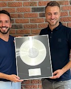 Dwóch uśmiechniętych mężczyzn prezentuje srebrną płytę z certyfikatem na tle ceglanej ściany. Jeden ubrany w niebieską koszulkę polo, drugi w niebieski t-shirt.