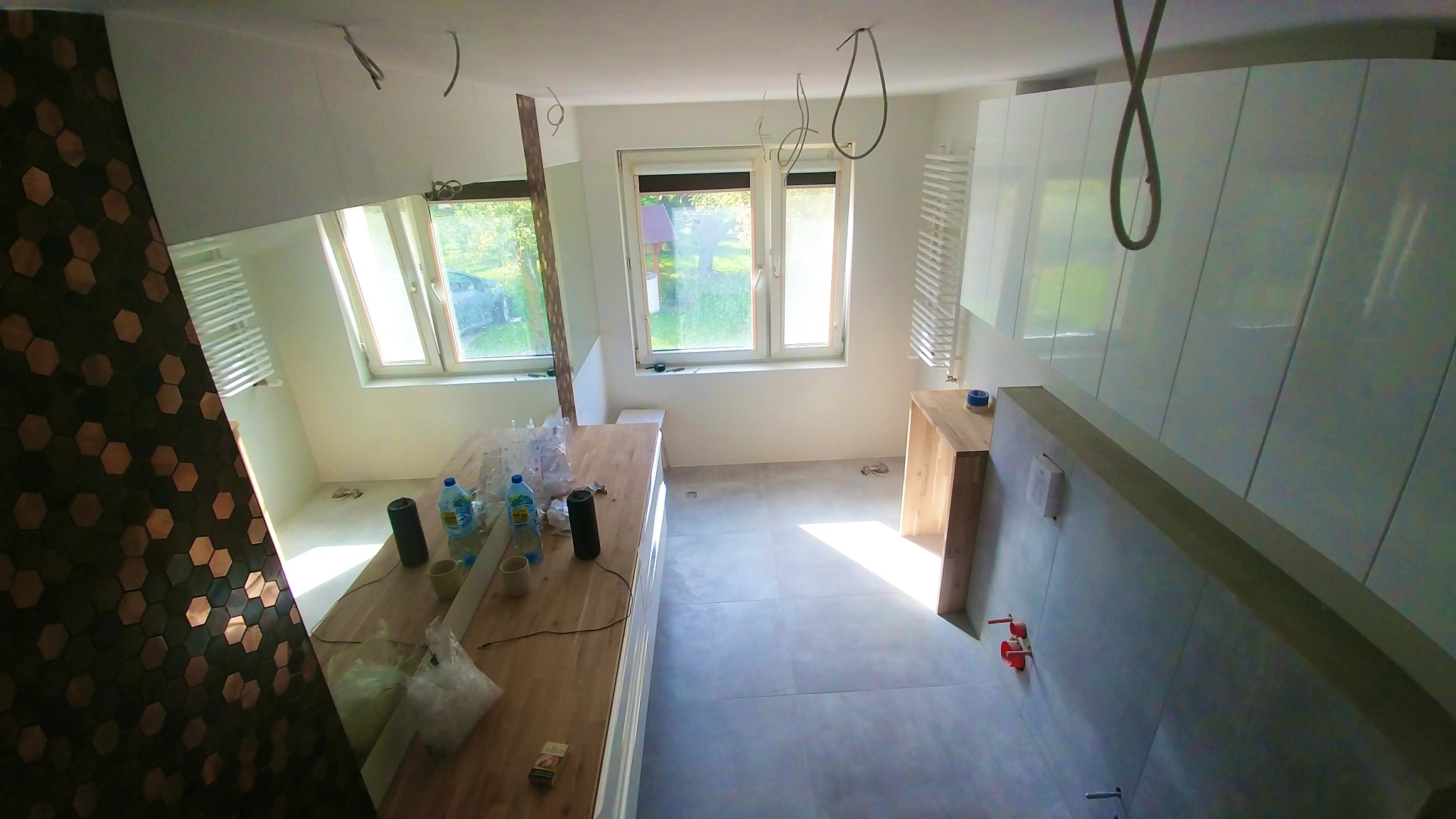 Wnętrze łazienki w trakcie wykańczania: widoczne płytki heksagonalne na ścianie, białe szafki, okna z widokiem na zieleń i instalacje. Częściowo widoczne materiały budowlane.