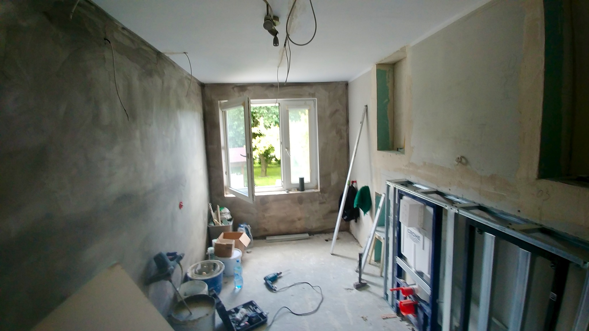 Pokój w trakcie prac wykończeniowych: surowe ściany, otwarte okno z widokiem na zieleń, narzędzia i materiały budowlane. Widoczna konstrukcja pod zabudowę.