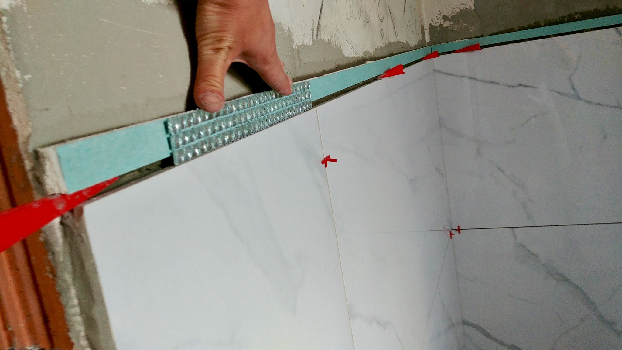 Układanie białych płytek z wzorem marmuru na ścianie, z użyciem poziomicy i czerwonych krzyżyków dystansowych. Widoczna dłoń glazurnika i fragment surowej ściany.