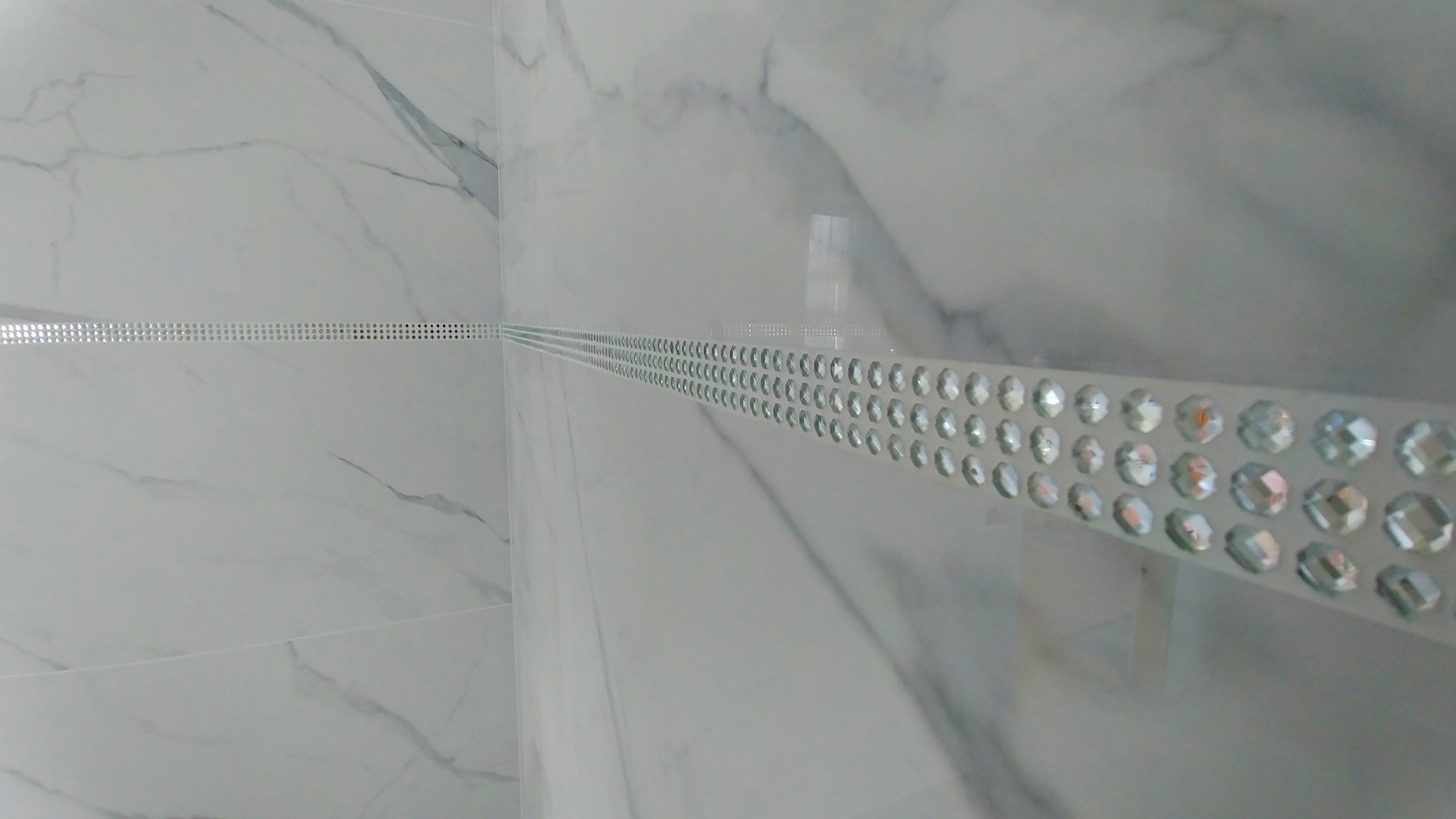 Białe marmurowe płytki z dekoracyjnym, błyszczącym paskiem z kryształkami, ułożone w łazience. Detal wykończenia wnętrza z bliska.
