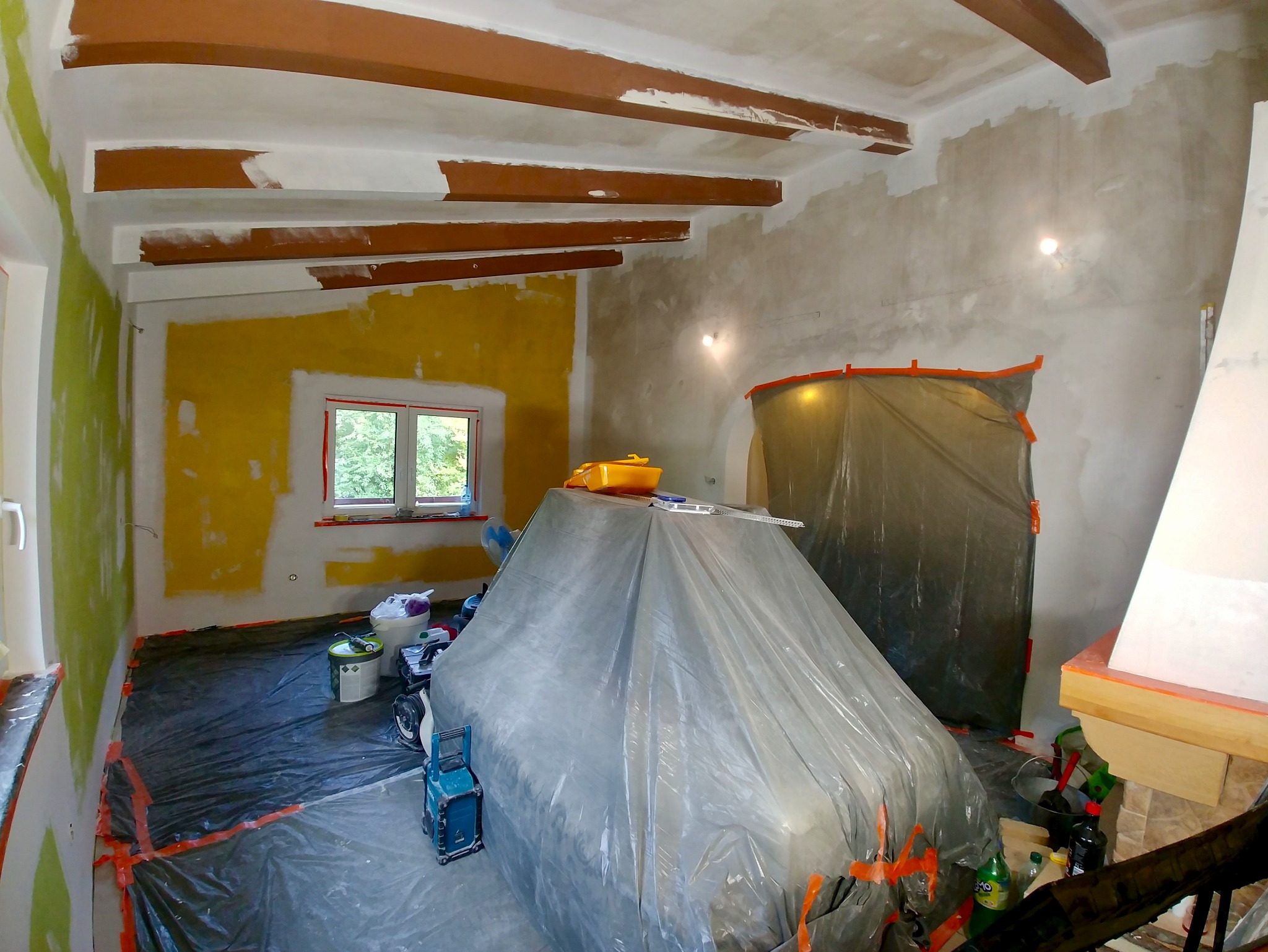 Pokój w trakcie malowania: ściany częściowo pokryte farbą, zabezpieczone meble folią, widoczne narzędzia malarskie i surowe drewniane belki na suficie.