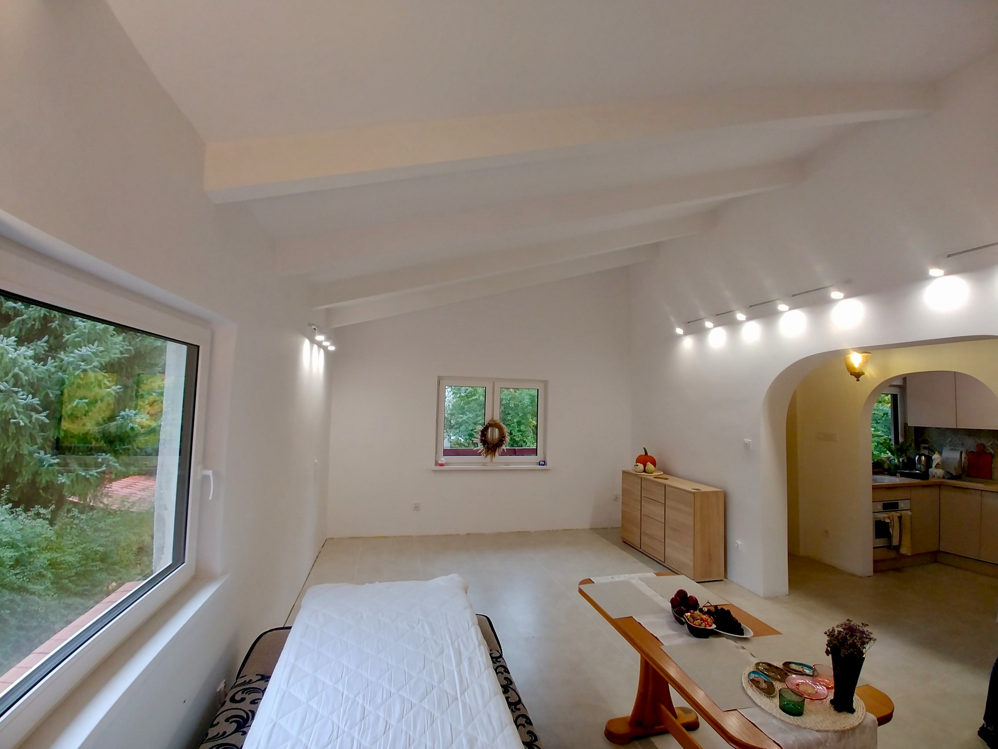 Jasne, przestronne wnętrze z białymi ścianami, drewnianymi meblami i oknem z widokiem na zieleń. Widoczne oświetlenie sufitowe i dekoracje okienne. Minimalistyczny styl.