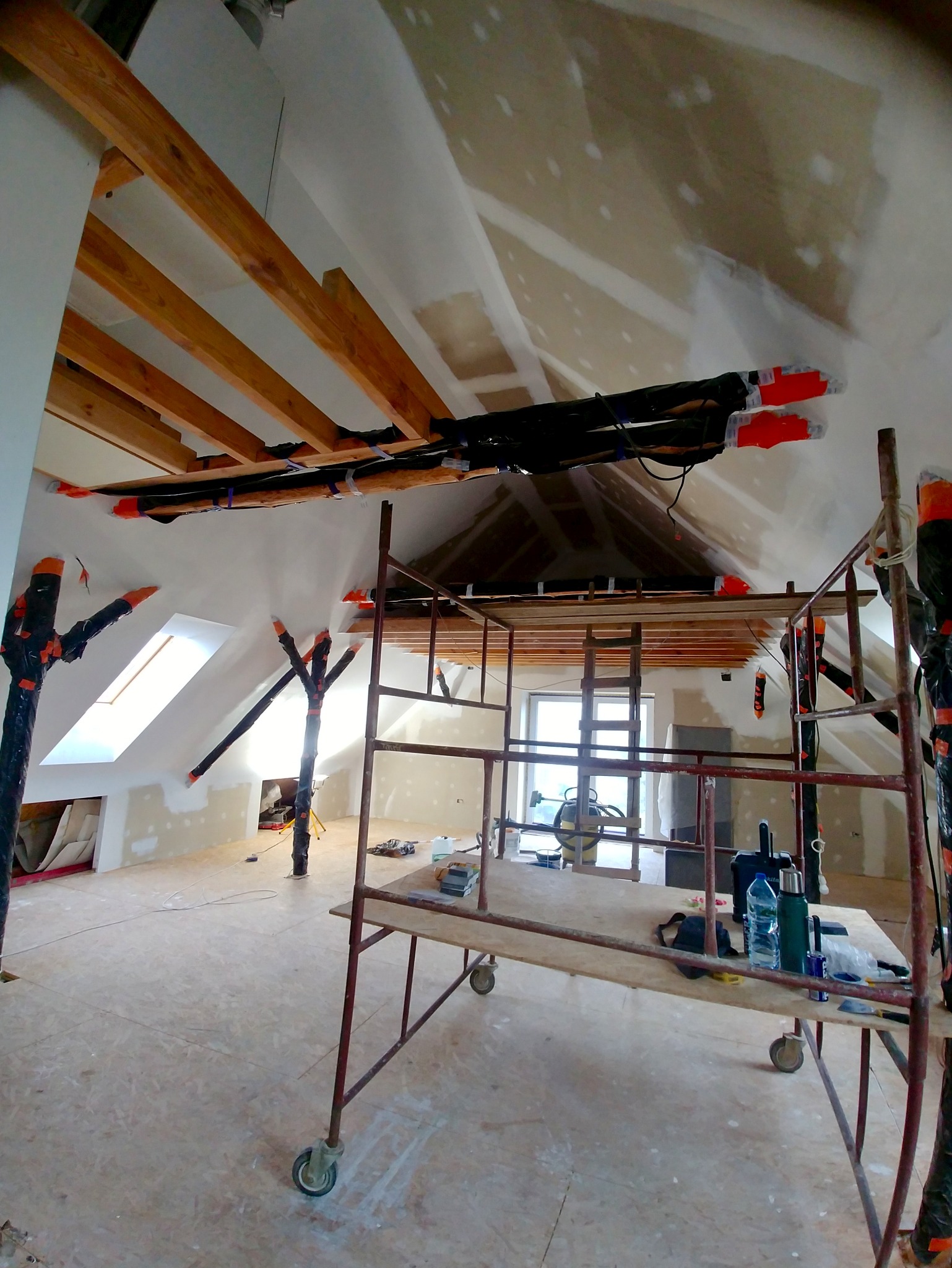 Poddasze w trakcie prac wykończeniowych: widoczne rusztowanie, ocieplenie rur, okna dachowe i przygotowane ściany do malowania. W tle widoczny odkurzacz przemysłowy.