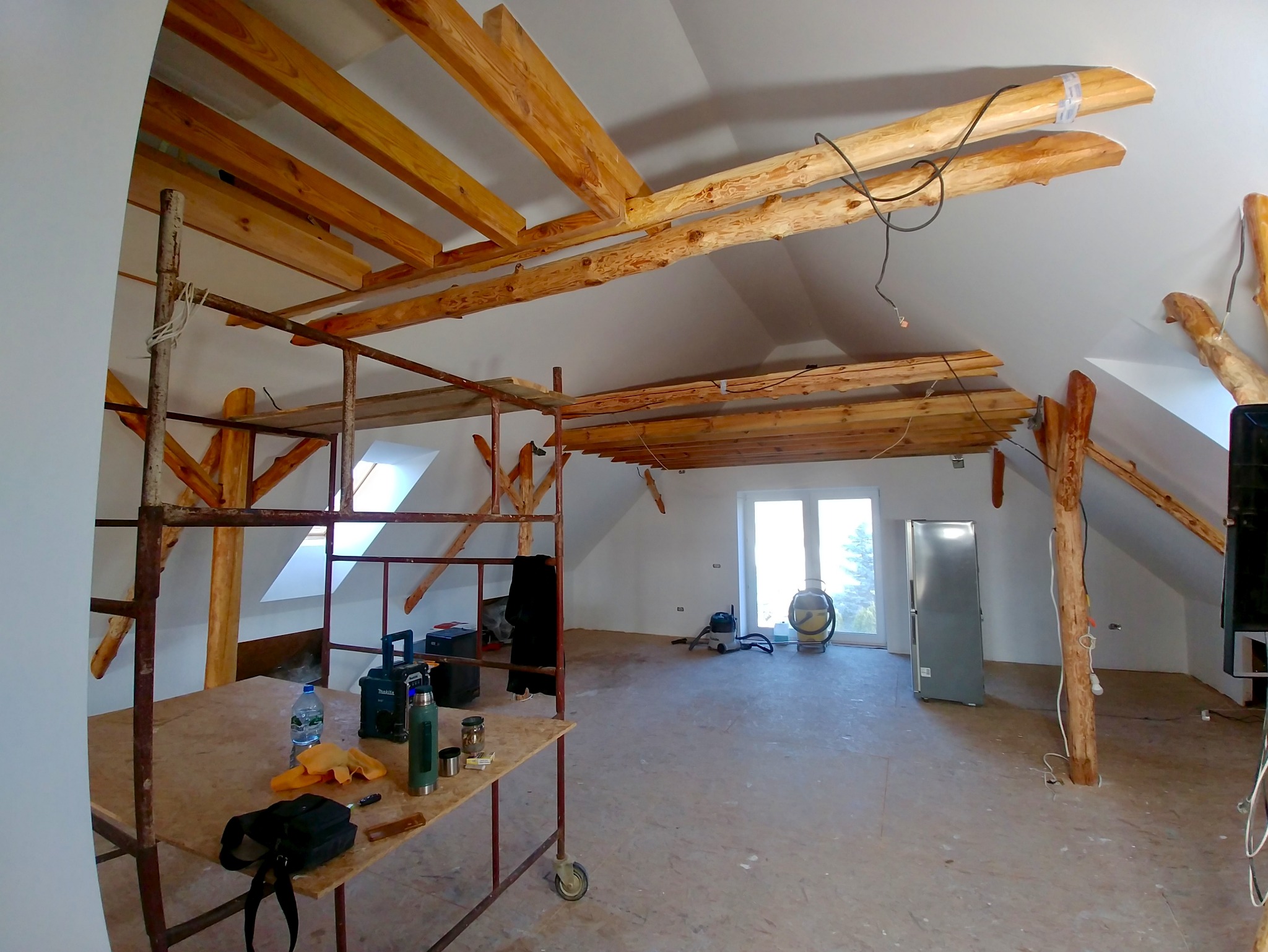 Poddasze w trakcie prac wykończeniowych z widocznym rusztowaniem, drewnianą konstrukcją dachu i oknami dachowymi. Na podłodze narzędzia i sprzęt budowlany.