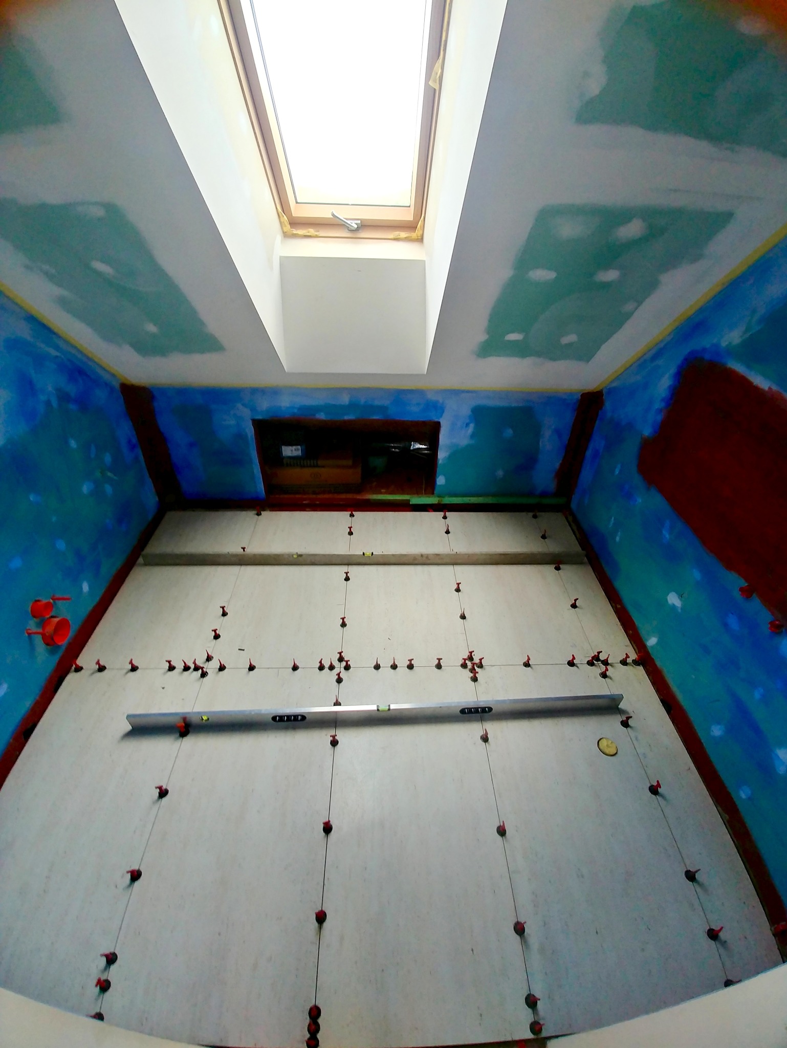 Ściana w trakcie układania płytek, poziomica, krzyżyki dystansowe, sufit z oknem dachowym, przygotowane ściany do wykończenia.