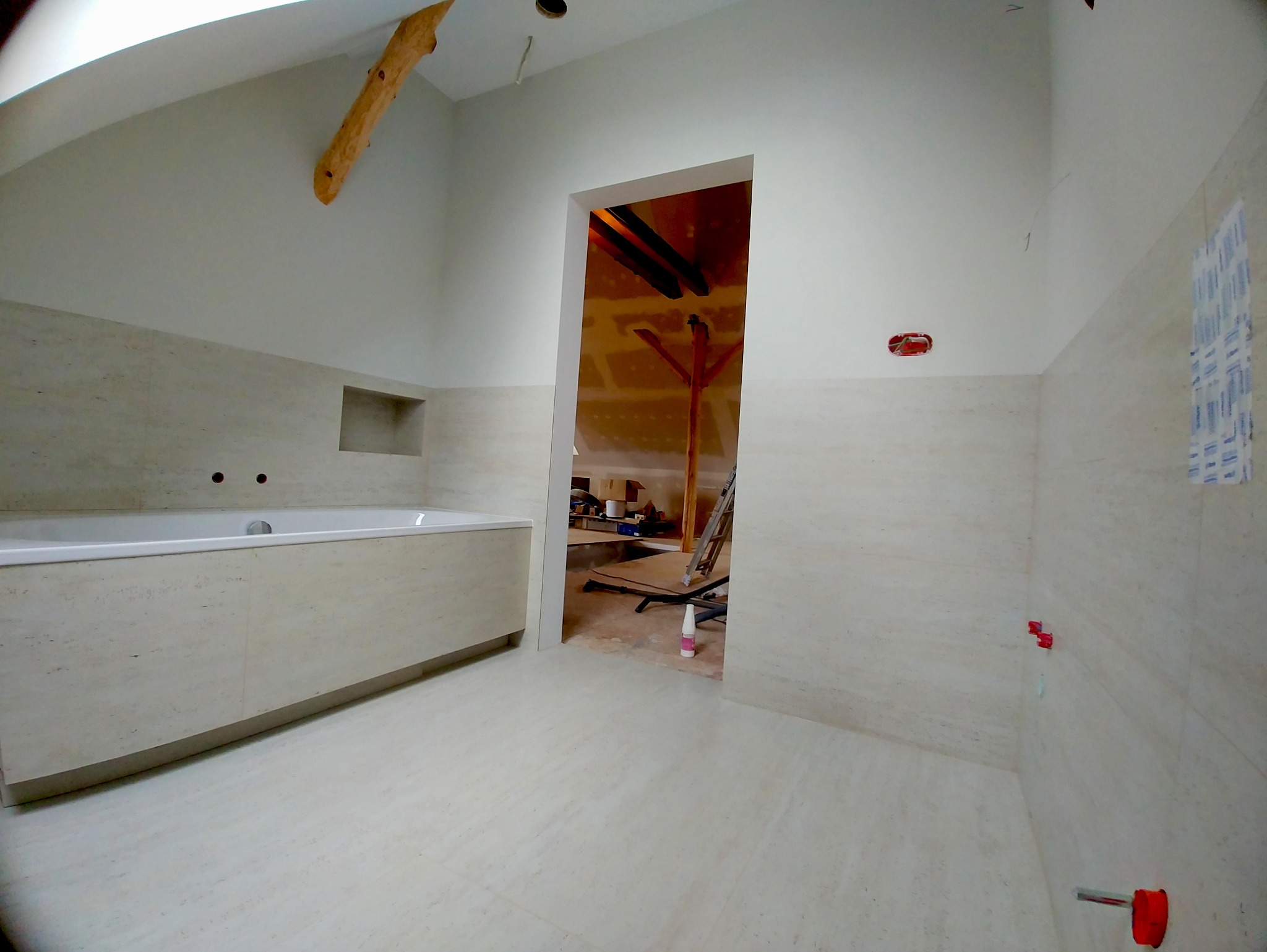 Jasna łazienka w trakcie wykańczania, widoczna wanna obudowana płytkami, fragment drewnianej belki stropowej i otwór drzwiowy do innego pomieszczenia.