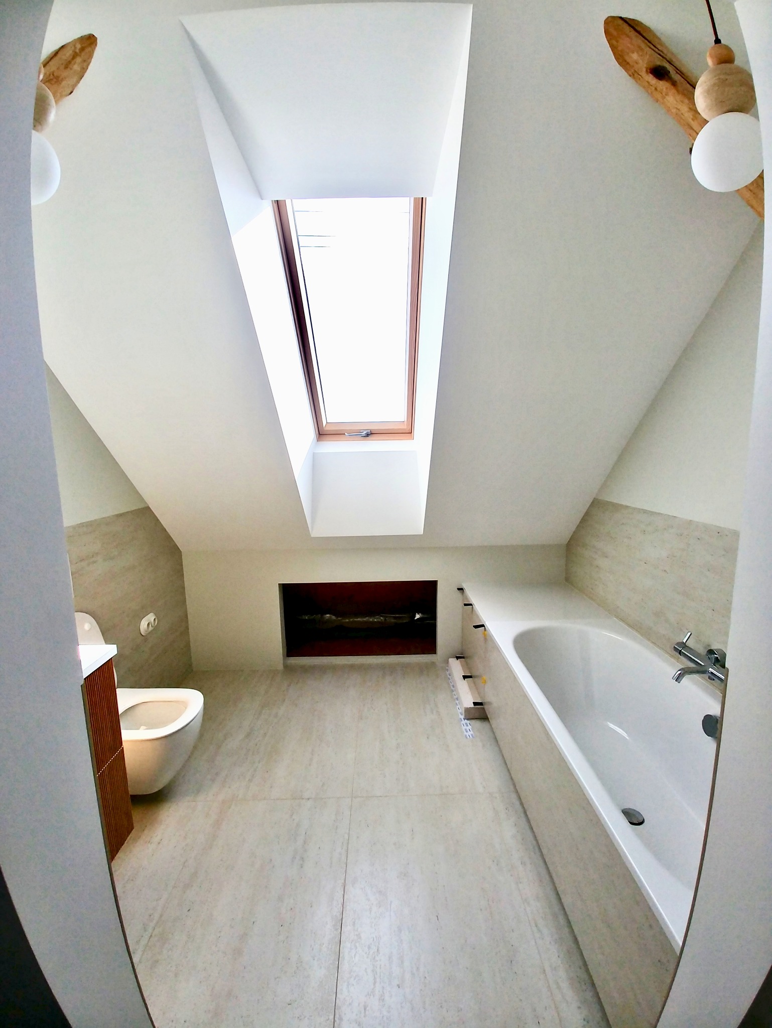 Jasna łazienka na poddaszu z oknem dachowym, beżowymi płytkami i wanną. Widoczne elementy dekoracyjne z drewna. Ujęcie z perspektywy wejścia do pomieszczenia.