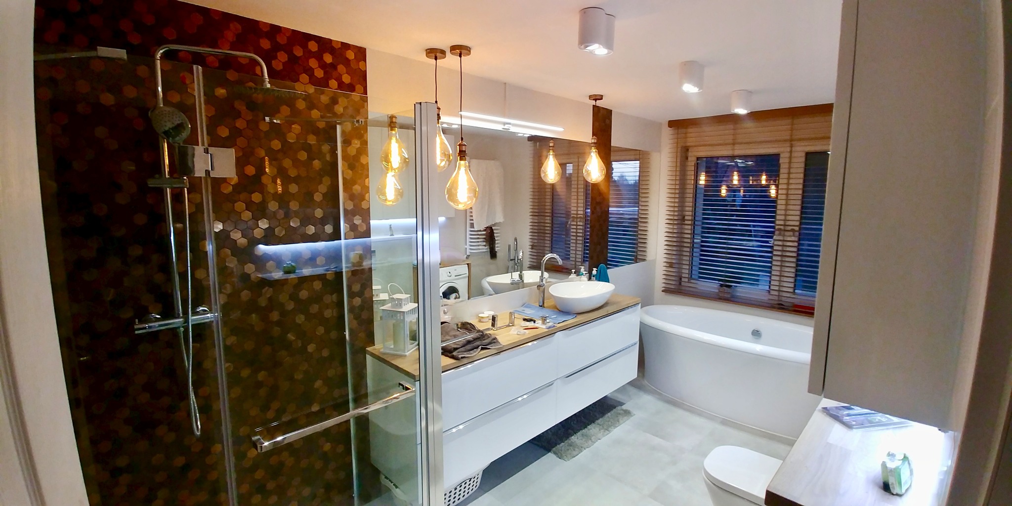 Nowoczesna łazienka z kabiną prysznicową wykończoną mozaiką heksagonalną, umywalką nablatową i oświetleniem dekoracyjnym. Widoczne okno z drewnianymi żaluzjami.