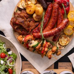 Bogaty zestaw grillowanych mięs, warzyw i sałatki na drewnianej desce, uzupełniony o różnorodne sosy w ceramicznych miseczkach. Idealny na imprezę plenerową.