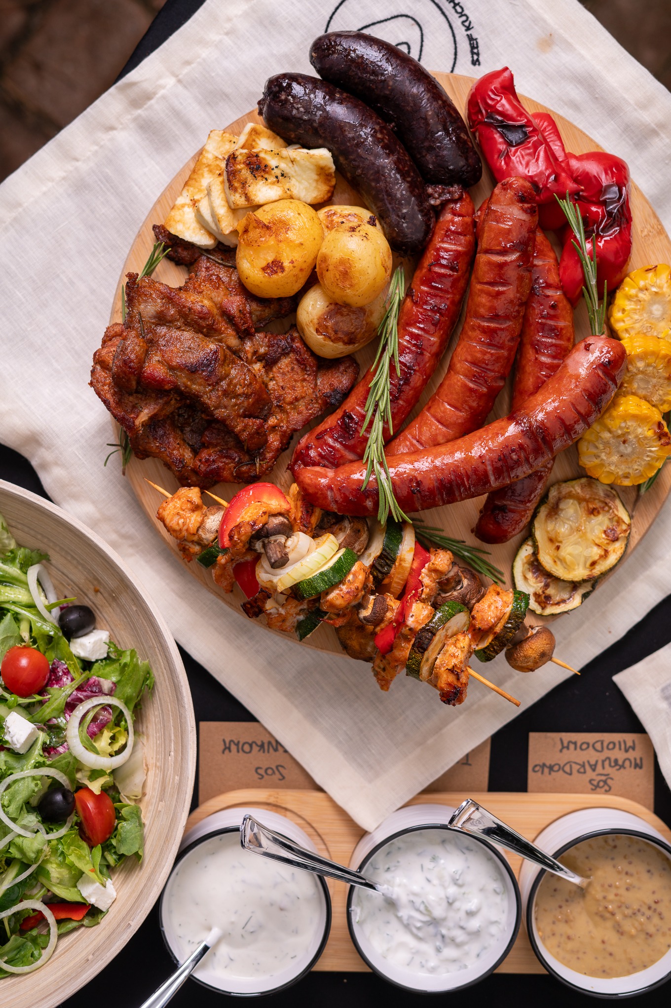 Bogaty zestaw grillowanych mięs, warzyw i sałatki na drewnianej desce, uzupełniony o różnorodne sosy w ceramicznych miseczkach. Idealny na imprezę plenerową.