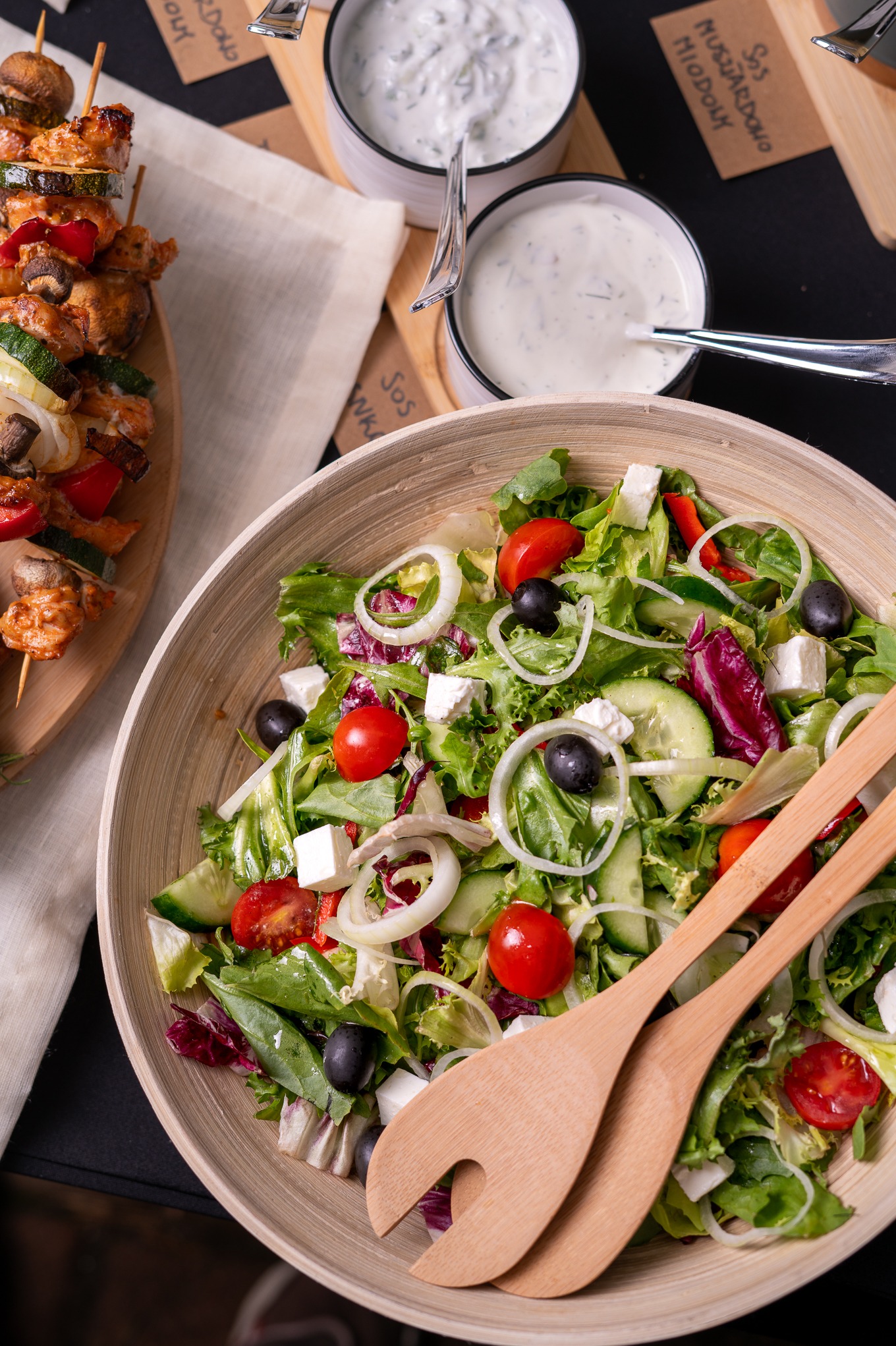 Bogata kompozycja kulinarna: szaszłyki z warzywami, miseczki z sosami i świeża sałatka grecka w drewnianej misie z drewnianymi sztućcami.