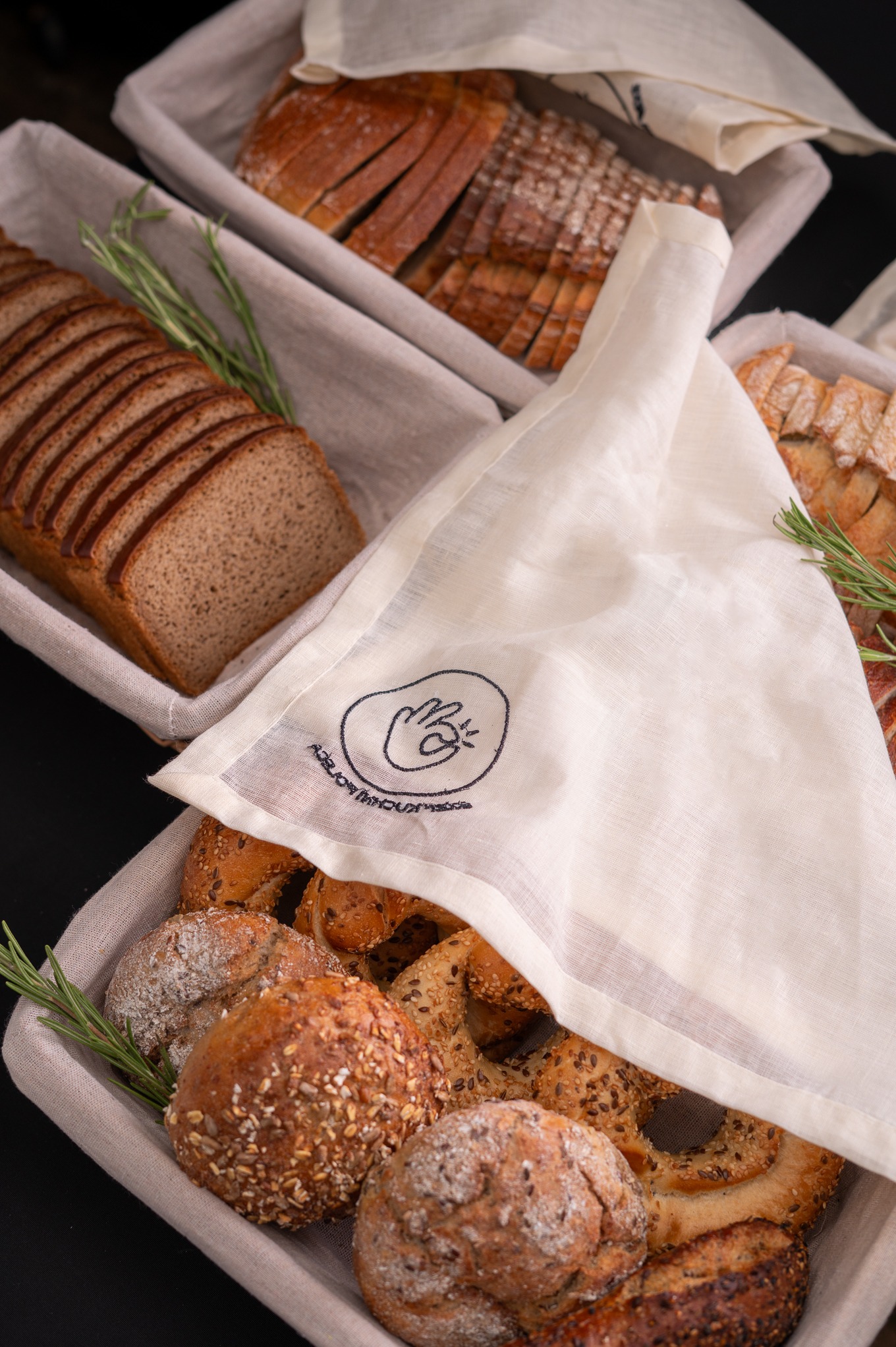 Kosz z pieczywem: chleb krojony, bułki posypane ziarnami, przykryte lnianą ściereczką z logo, gałązka rozmarynu. Prezentacja wyrobów piekarniczych.