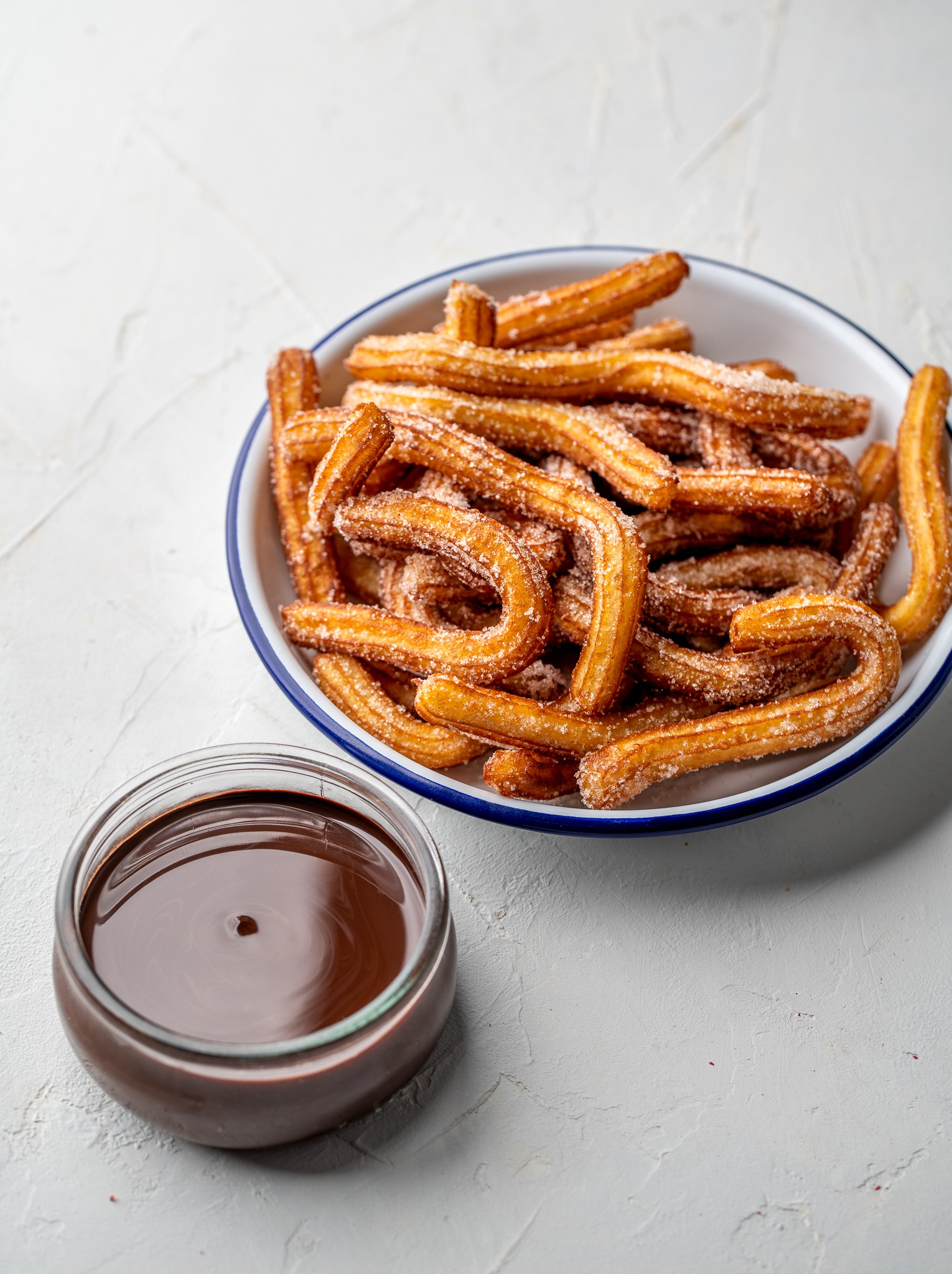 Churros posypane cukrem w emaliowanej misce z niebieskim rantem, obok szklana miseczka z sosem czekoladowym na białym teksturowanym tle.