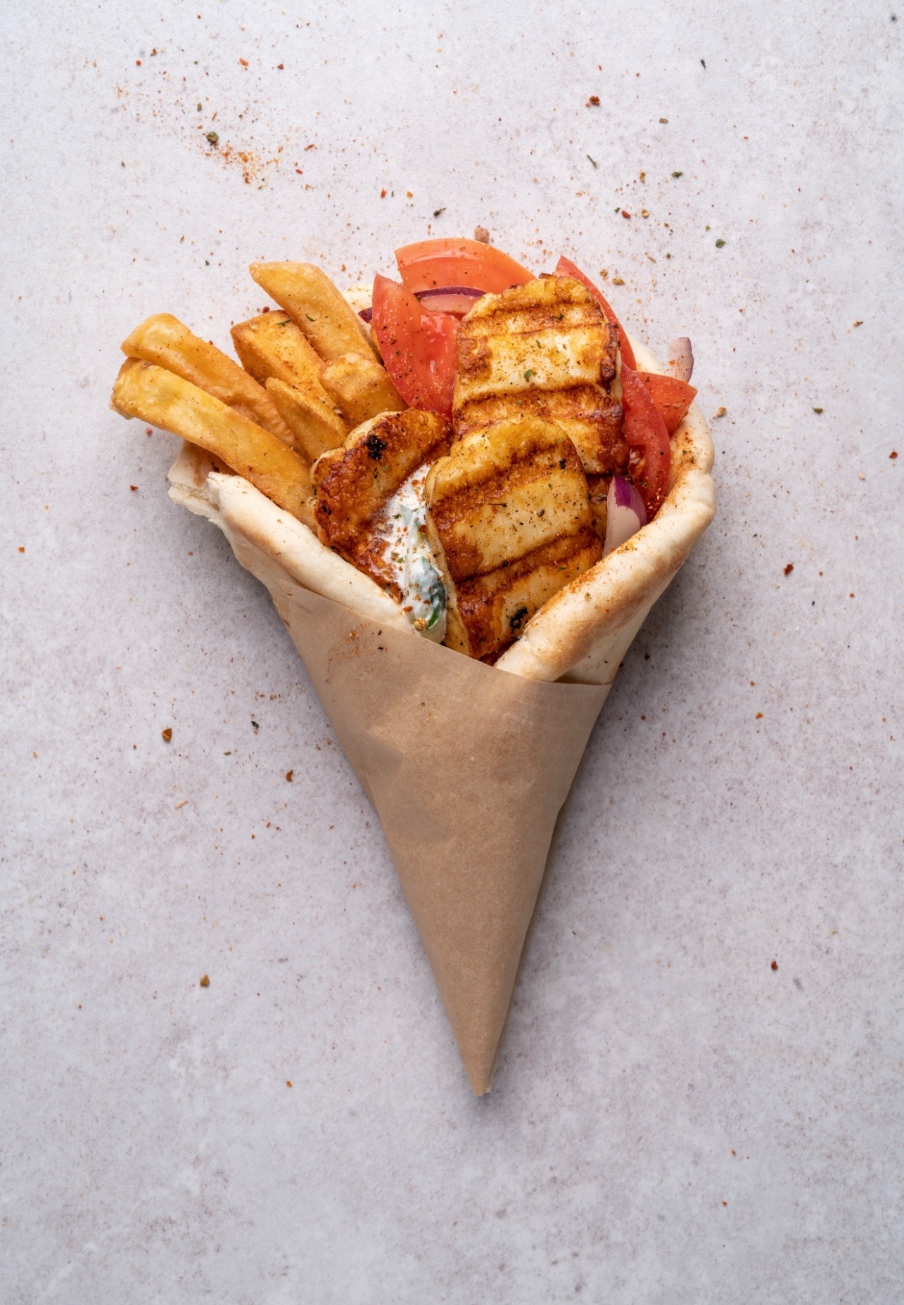 Pita z grillowanym serem halloumi, frytkami, pomidorami i sosem tzatziki, zawinięta w papierową torebkę, na jasnym tle z przyprawami. Widok z góry.