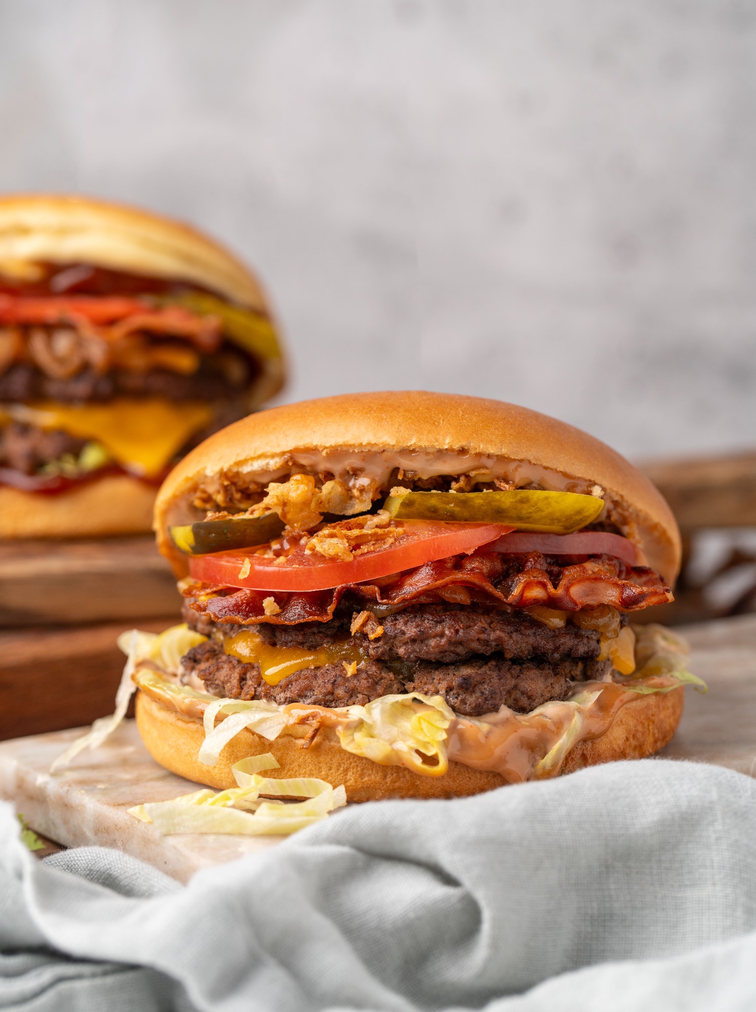 Apetyczny, dwuwarstwowy burger z bekonem, serem cheddar, pomidorem i piklami, prezentowany na drewnianej desce z lnianą ściereczką. W tle drugi burger.