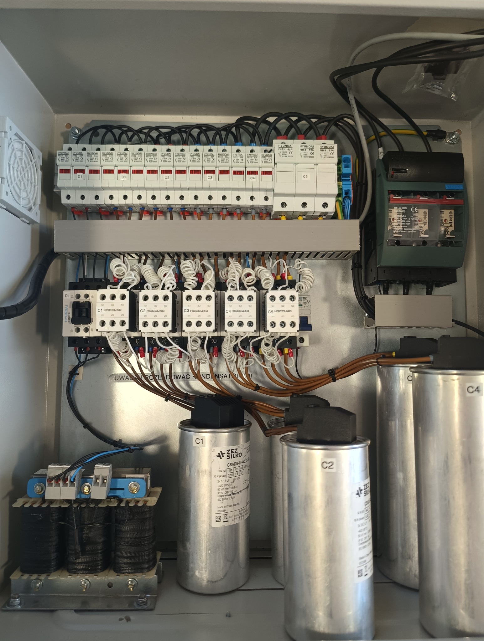 Rozdzielnica elektryczna z bezpiecznikami, stycznikami i kondensatorami w szarej obudowie. Widoczne przewody i oznaczenia elementów.