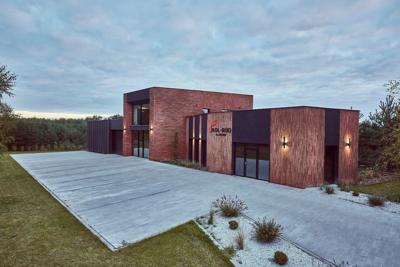 Nowoczesny budynek biurowy z elewacją z cegły i drewna, oświetlony lampami ściennymi, z dużym parkingiem z kostki brukowej i zielonym trawnikiem.