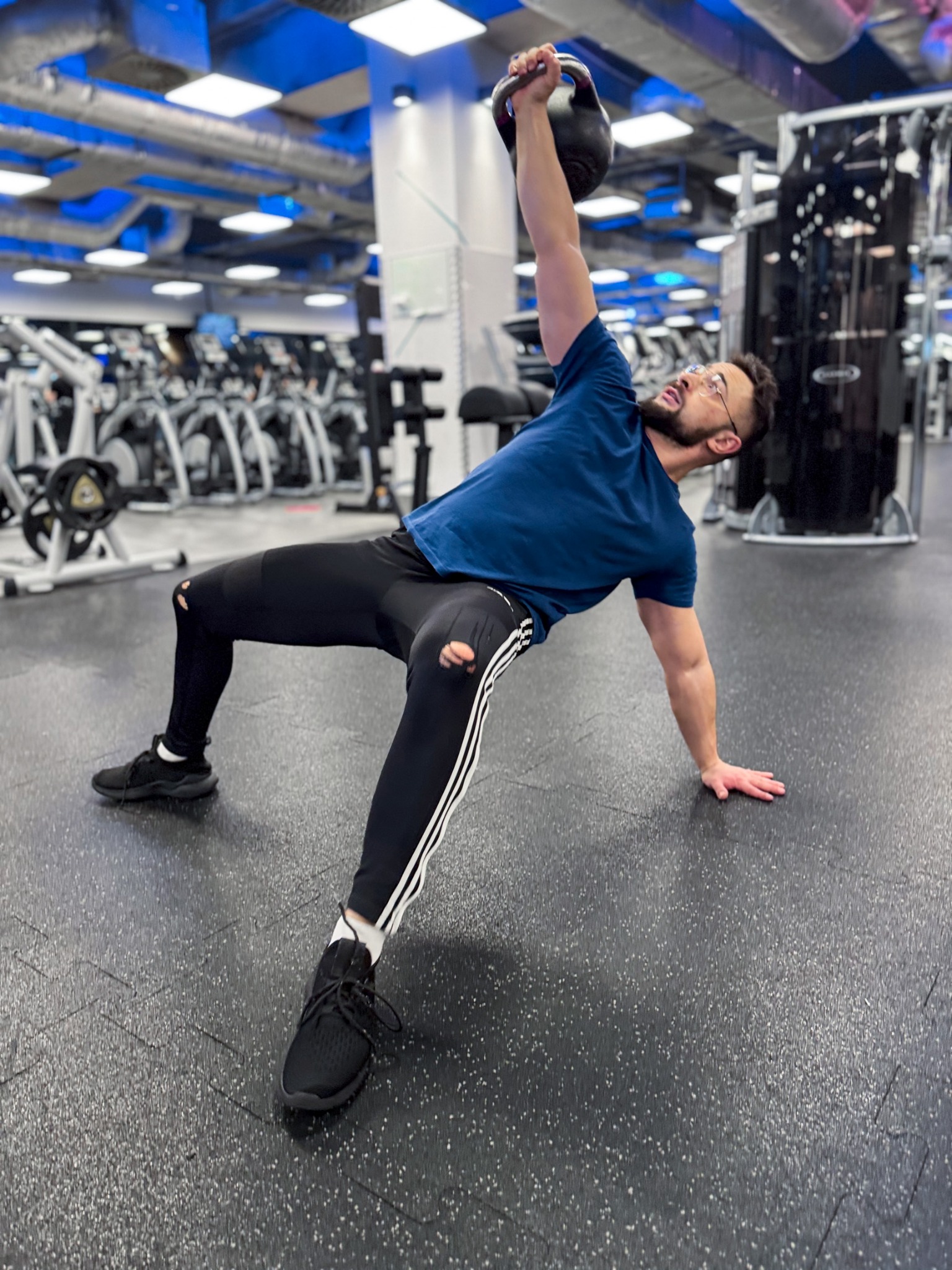 Mężczyzna w sportowym stroju, z brodą i okularach, wykonuje ćwiczenie z kettlebell w siłowni, opierając się na jednej ręce, w tle sprzęt do ćwiczeń.