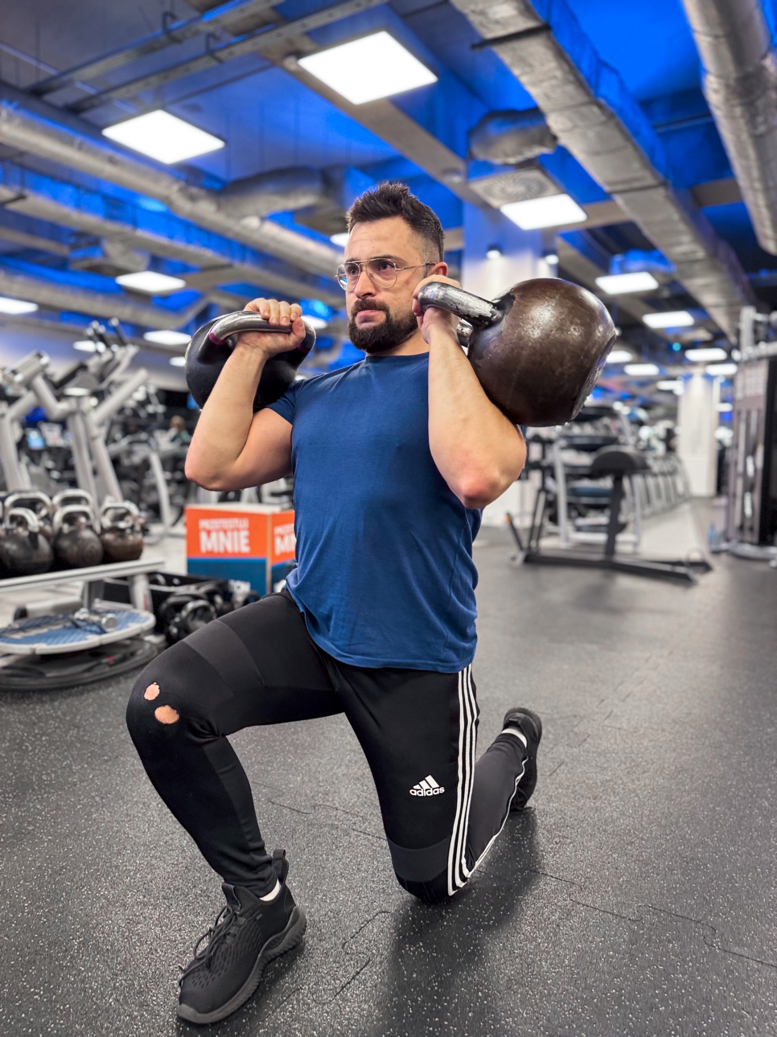 Mężczyzna w okularach klęczy w siłowni, trzymając dwie kule kettlebell na ramionach. Widoczne podarte legginsy i sprzęt do ćwiczeń w tle.