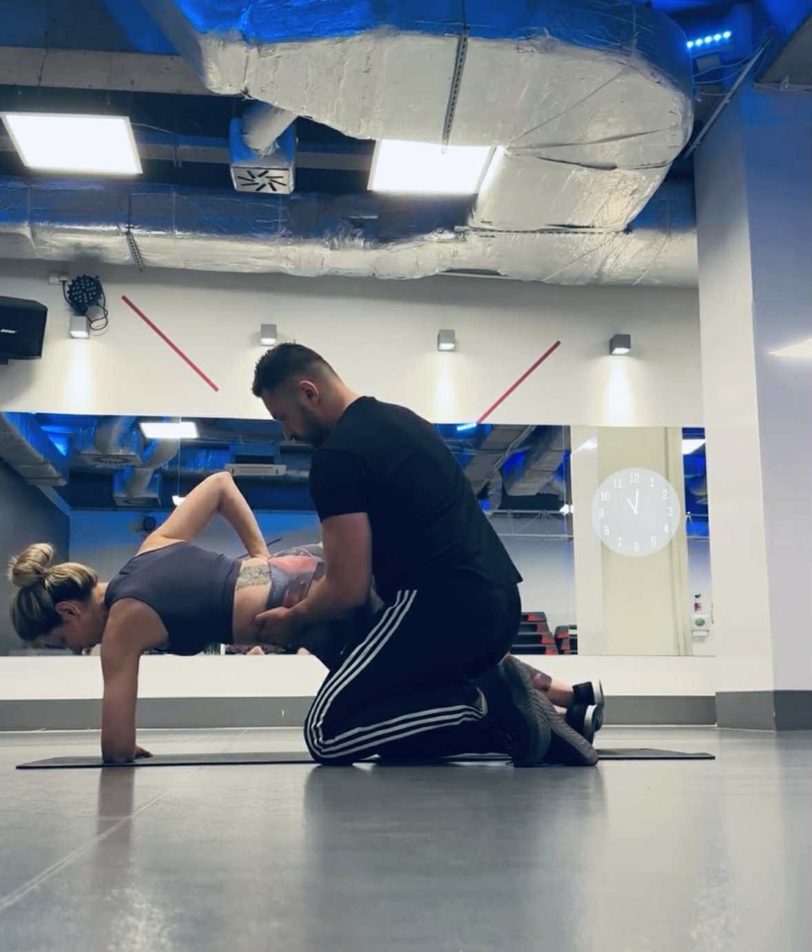 Trener personalny koryguje pozycję deski kobiety na macie w siłowni. Widoczna duża przestrzeń, lustra i instalacje na suficie.