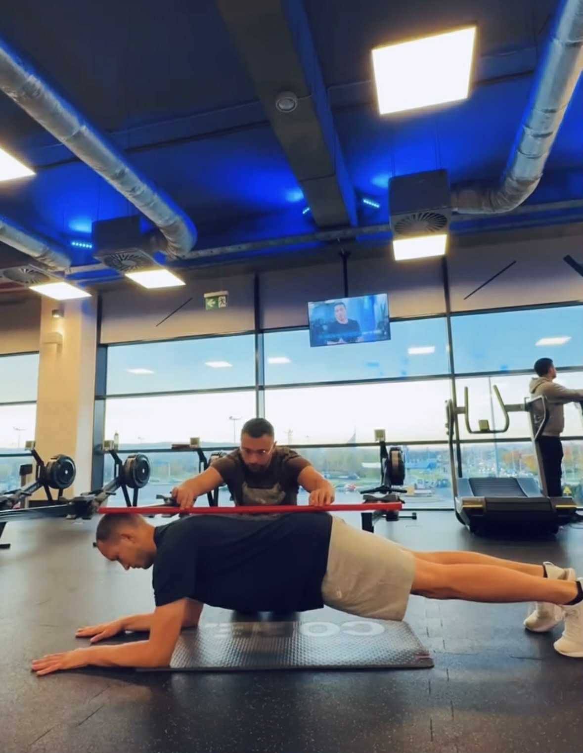 Trener koryguje pozycję ćwiczącego podczas deski w jasnej siłowni z dużymi oknami i widocznym sprzętem fitness w tle. Ćwiczenie z użyciem czerwonej laski.