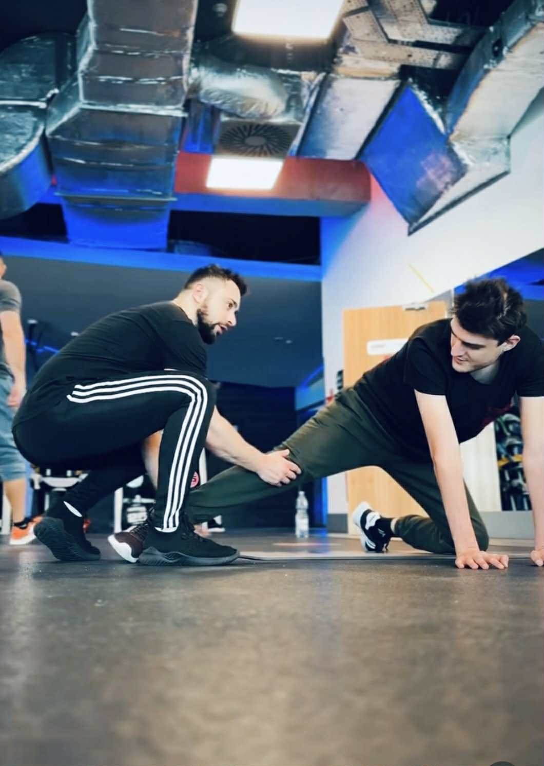 Osobisty instruktor w czarnym stroju pomaga mężczyźnie w rozciąganiu nogi na podłodze w siłowni z widocznymi rurami wentylacyjnymi pod sufitem.