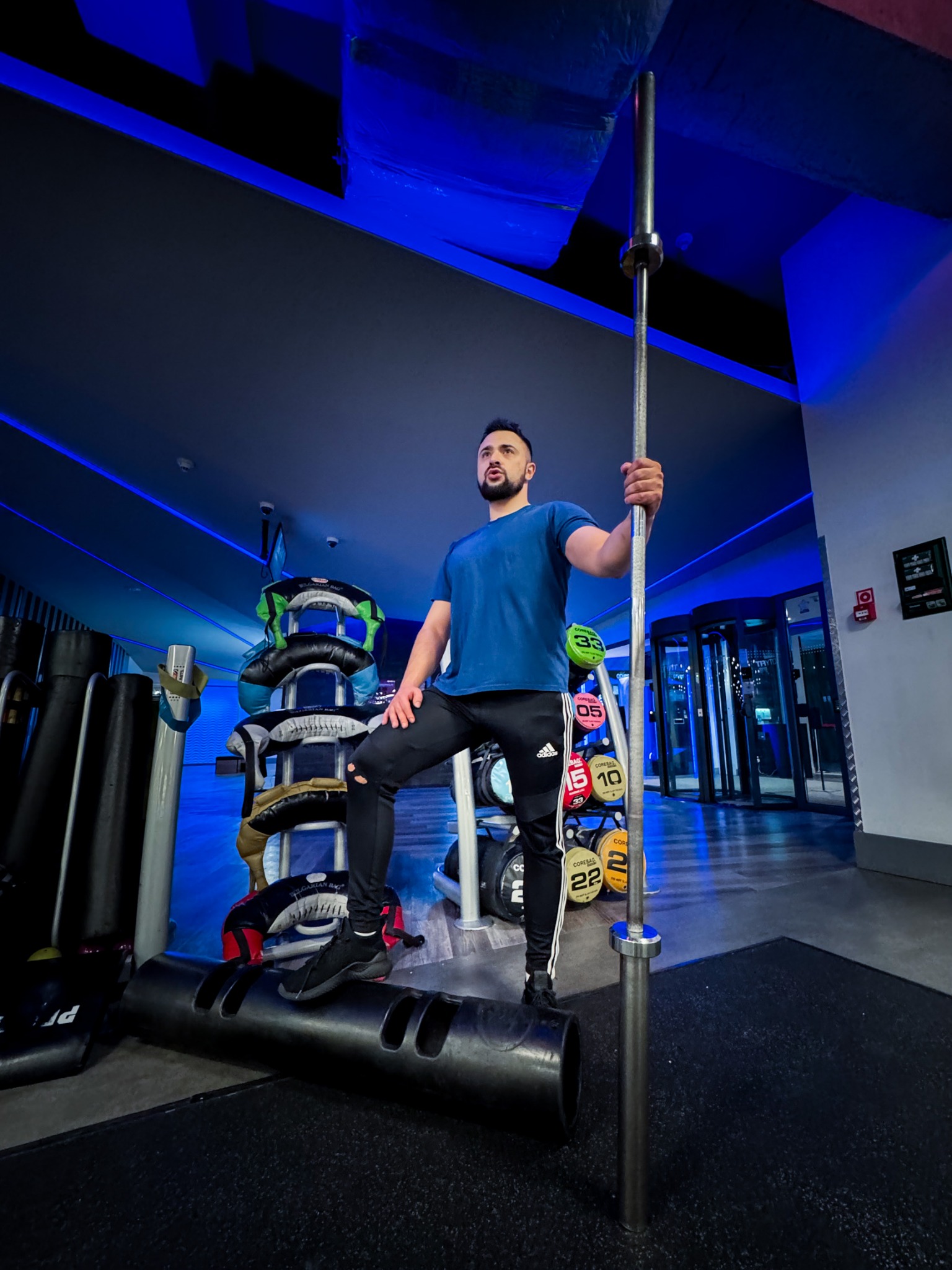 Mężczyzna w sportowym stroju, z uniesioną sztangą, pozuje w nowoczesnej siłowni z niebieskim oświetleniem i sprzętem do ćwiczeń. Dynamiczna poza.