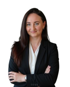 Portret kobiety w czarnej marynarce na białym tle, z rękami skrzyżowanymi na piersi. Delikatny uśmiech, ciemne włosy opadają na ramiona. Elegancki zegarek na nadgarstku.