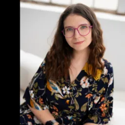 Portret kobiety w kwiecistej sukience i różowych okularach, siedzącej na białej sofie w jasnym pomieszczeniu. Uśmiech, naturalne światło, relaksująca atmosfera.