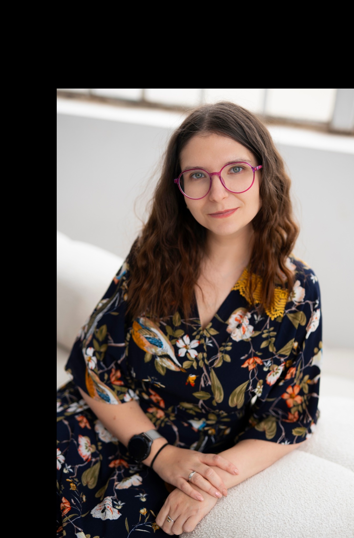 Portret kobiety w kwiecistej sukience i różowych okularach, siedzącej na białej sofie w jasnym pomieszczeniu. Uśmiech, naturalne światło, relaksująca atmosfera.