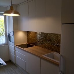 DWÓCH OD MEBLI Artur Sikorzyński - Biała kuchnia z drewnianym blatem, złotą mozaiką na ścianie i wiszącą lampą w stylu retro. Widoczne okno z zasłonką i fragment podłogi z paneli.