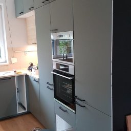 DWÓCH OD MEBLI Artur Sikorzyński - Szara zabudowa kuchenna z czarnymi uchwytami i wbudowanym piekarnikiem, widoczny stołek i częściowo otwarte szafki. Ujęcie z boku prezentuje montaż.