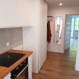 DWÓCH OD MEBLI Artur Sikorzyński - Nowoczesna kuchnia z białymi frontami, drewnianym blatem i zabudowaną szafą w przedpokoju, widok z perspektywy wejścia, podłoga z paneli.