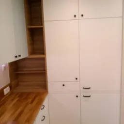 Biała zabudowa kuchenna z drewnianym blatem i otwartymi półkami z naturalnego drewna. Minimalistyczne, czarne uchwyty. Widok na narożnik kuchni.