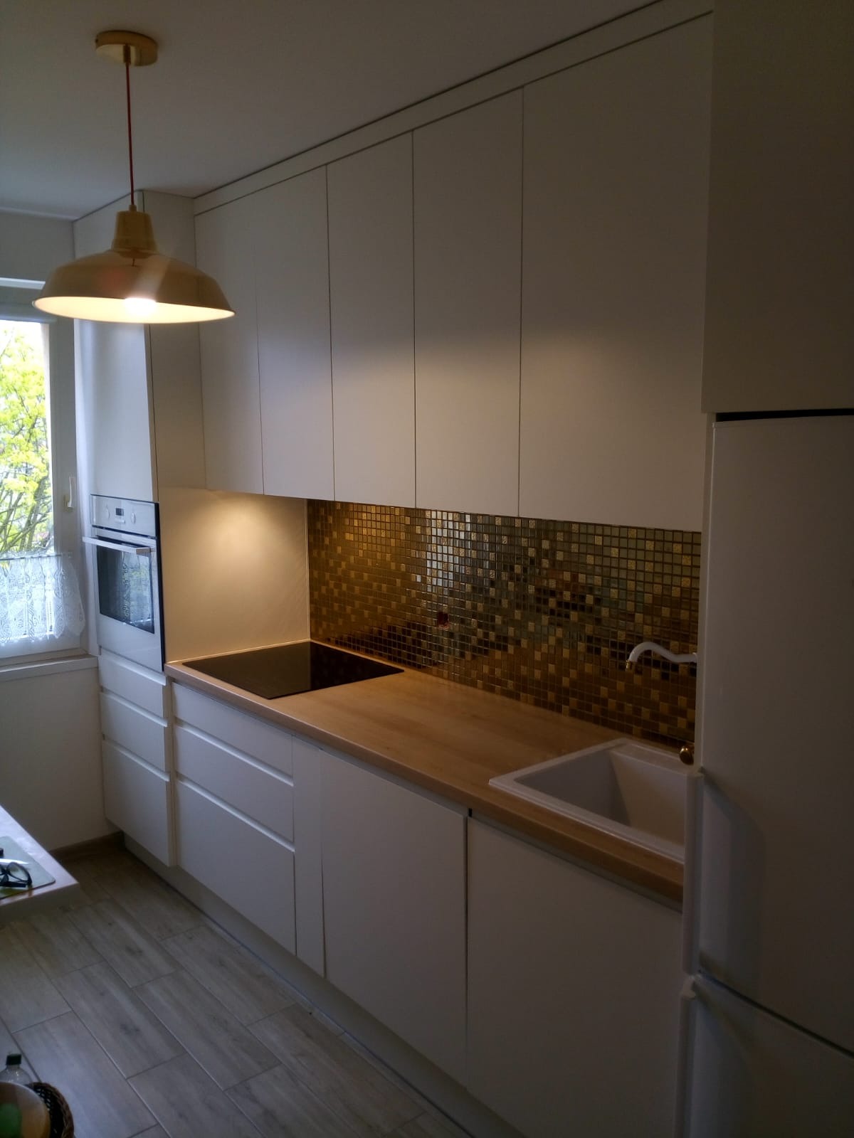 Biała kuchnia z drewnianym blatem, złotą mozaiką na ścianie i wiszącą lampą w stylu retro. Widoczne okno z zasłonką i fragment podłogi z paneli.
