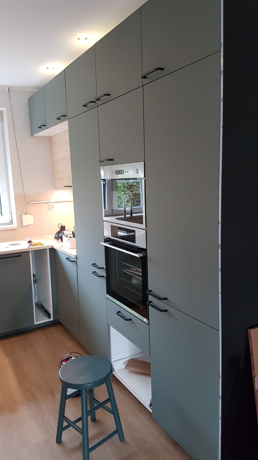 Szara zabudowa kuchenna z czarnymi uchwytami i wbudowanym piekarnikiem, widoczny stołek i częściowo otwarte szafki. Ujęcie z boku prezentuje montaż.
