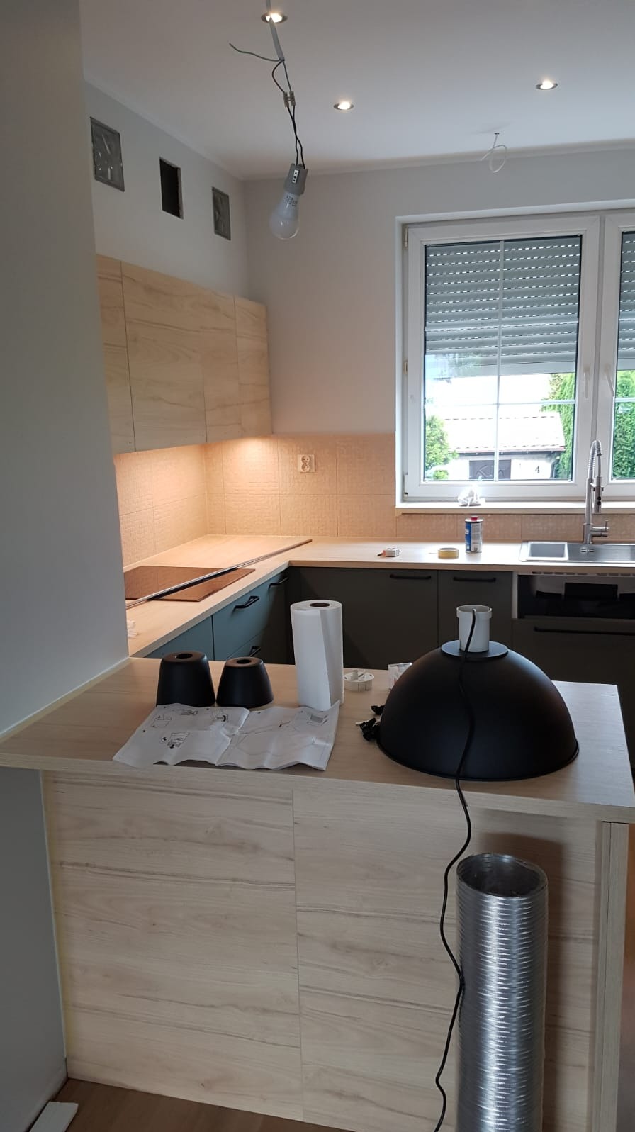 Jasna kuchnia z szarymi szafkami i drewnianym blatem. Na pierwszym planie elementy oświetlenia i instrukcja montażu, sugerujące etap wykończeniowy.