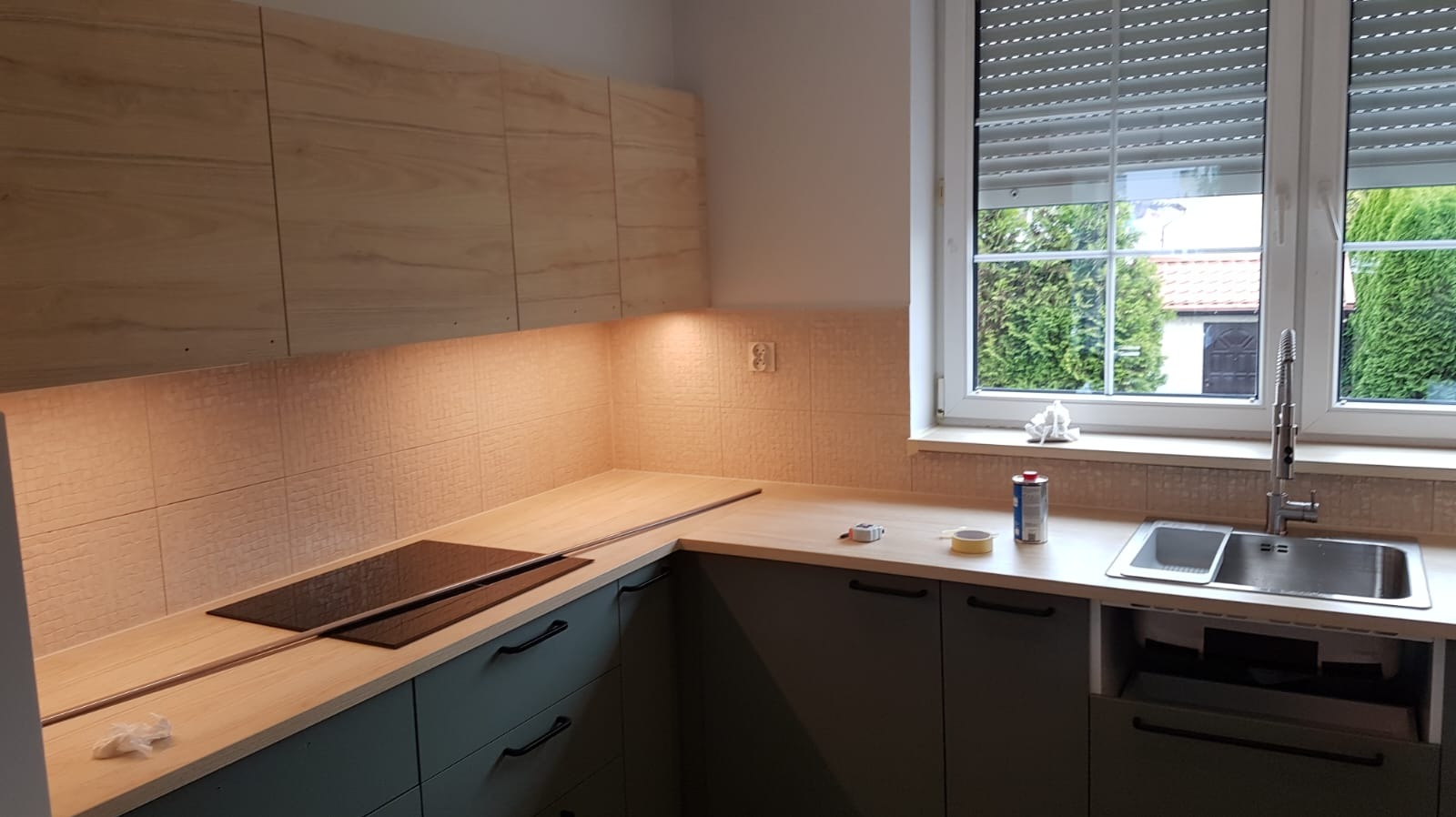 Nowoczesna kuchnia z drewnianymi szafkami górnymi i szarymi dolnymi, z wbudowaną płytą grzewczą i oświetleniem podszafkowym, widok na okno z roletami.