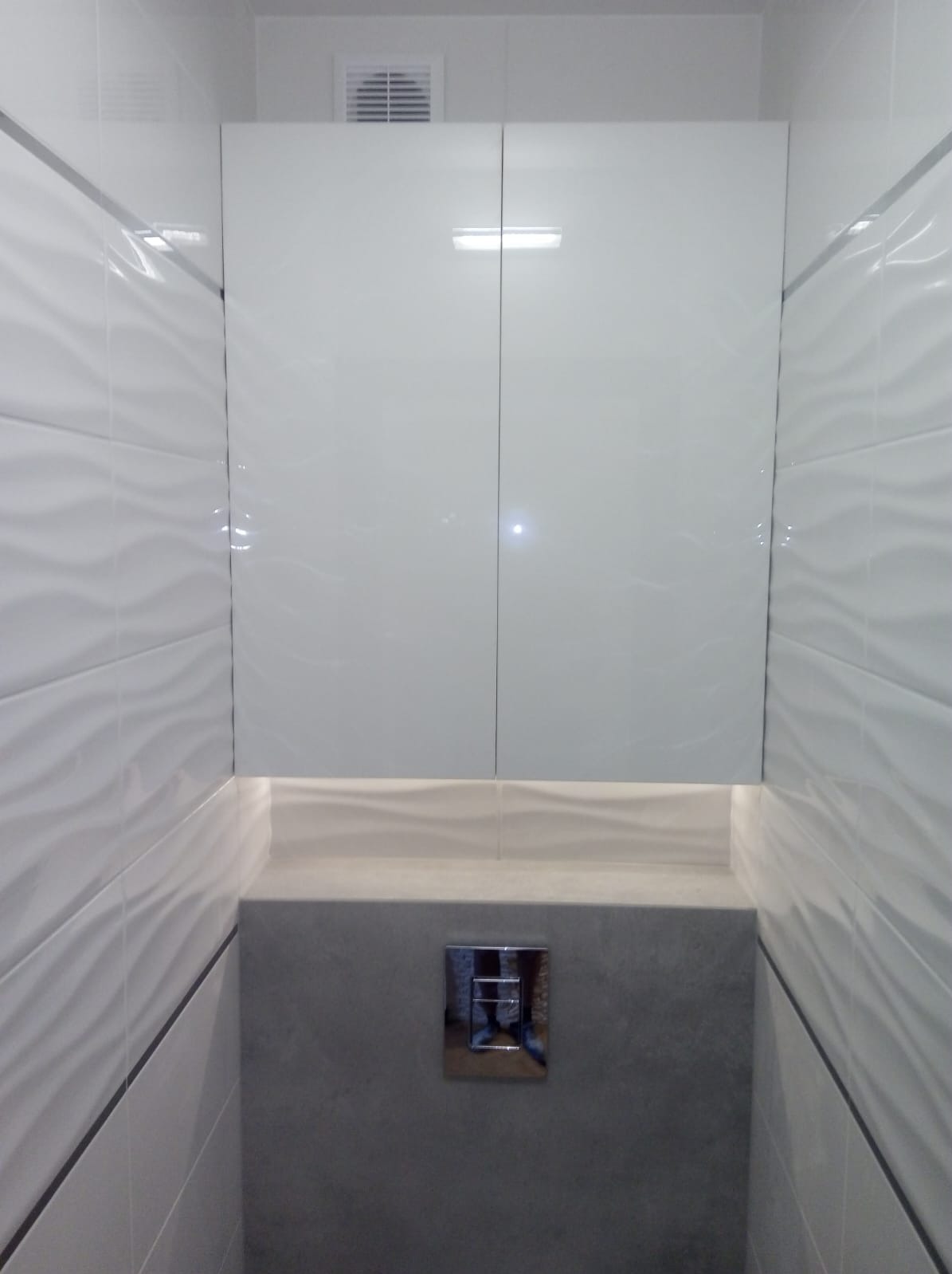 Nowoczesna, biała szafka wnękowa z połyskiem, zamontowana nad toaletą. Wzór płytek imituje fale. Chromowany przycisk spłuczki. Minimalistyczny design.