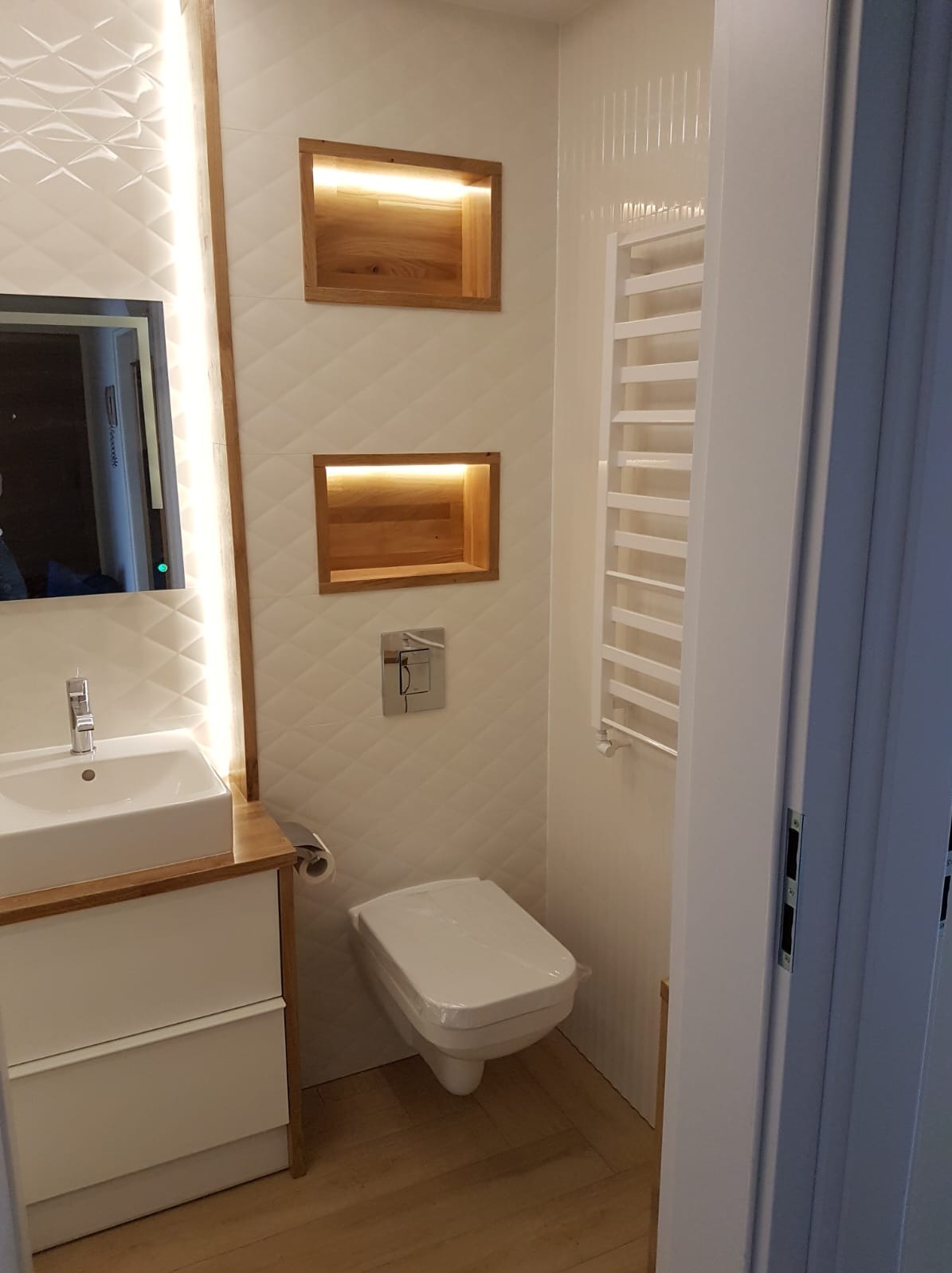 Nowoczesna łazienka z białymi płytkami 3D, drewnianymi wnękami z oświetleniem, umywalką z szafką i grzejnikiem drabinkowym. Widok zza uchylonych drzwi.