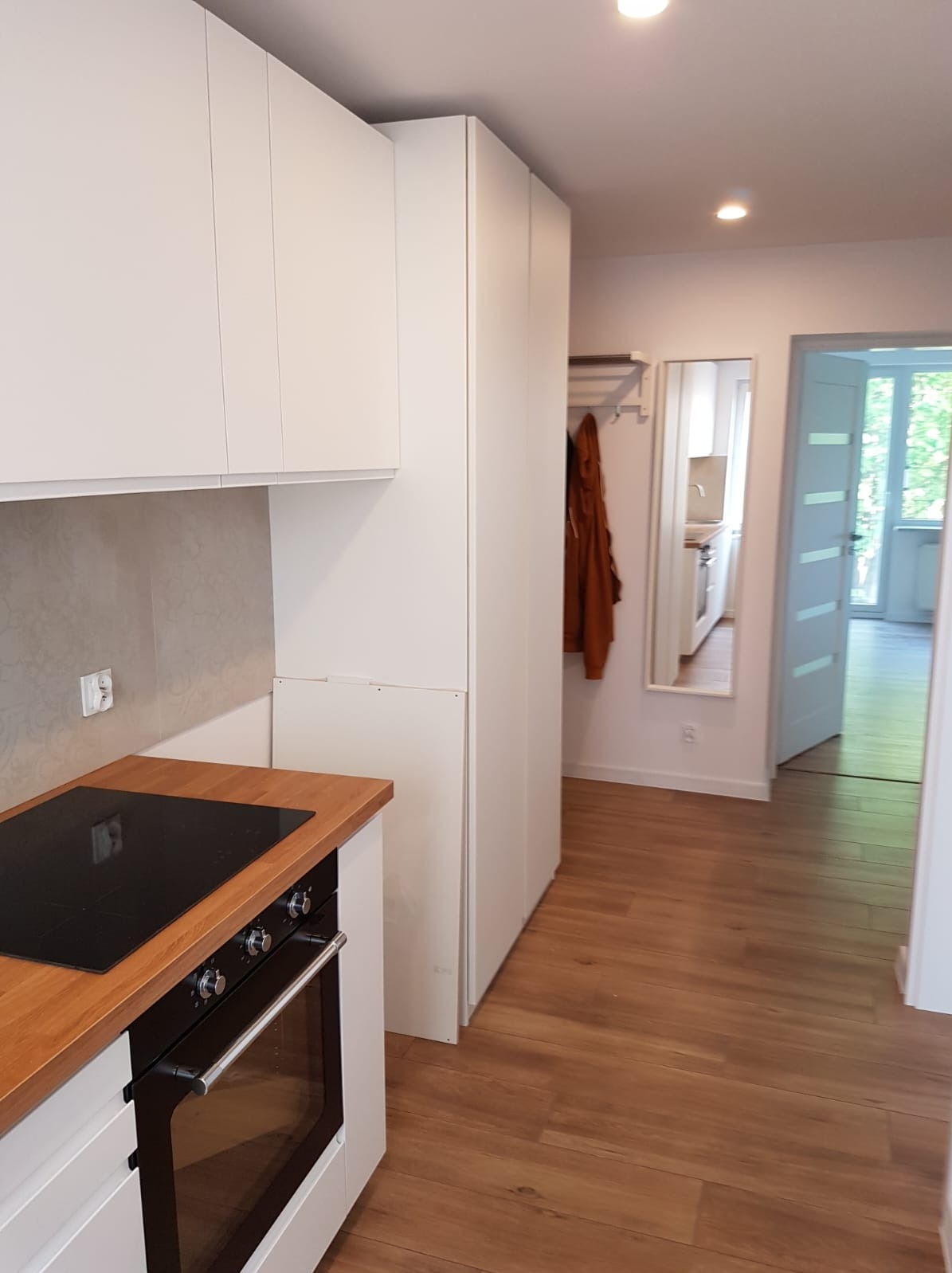 Nowoczesna kuchnia z białymi frontami, drewnianym blatem i zabudowaną szafą w przedpokoju, widok z perspektywy wejścia, podłoga z paneli.