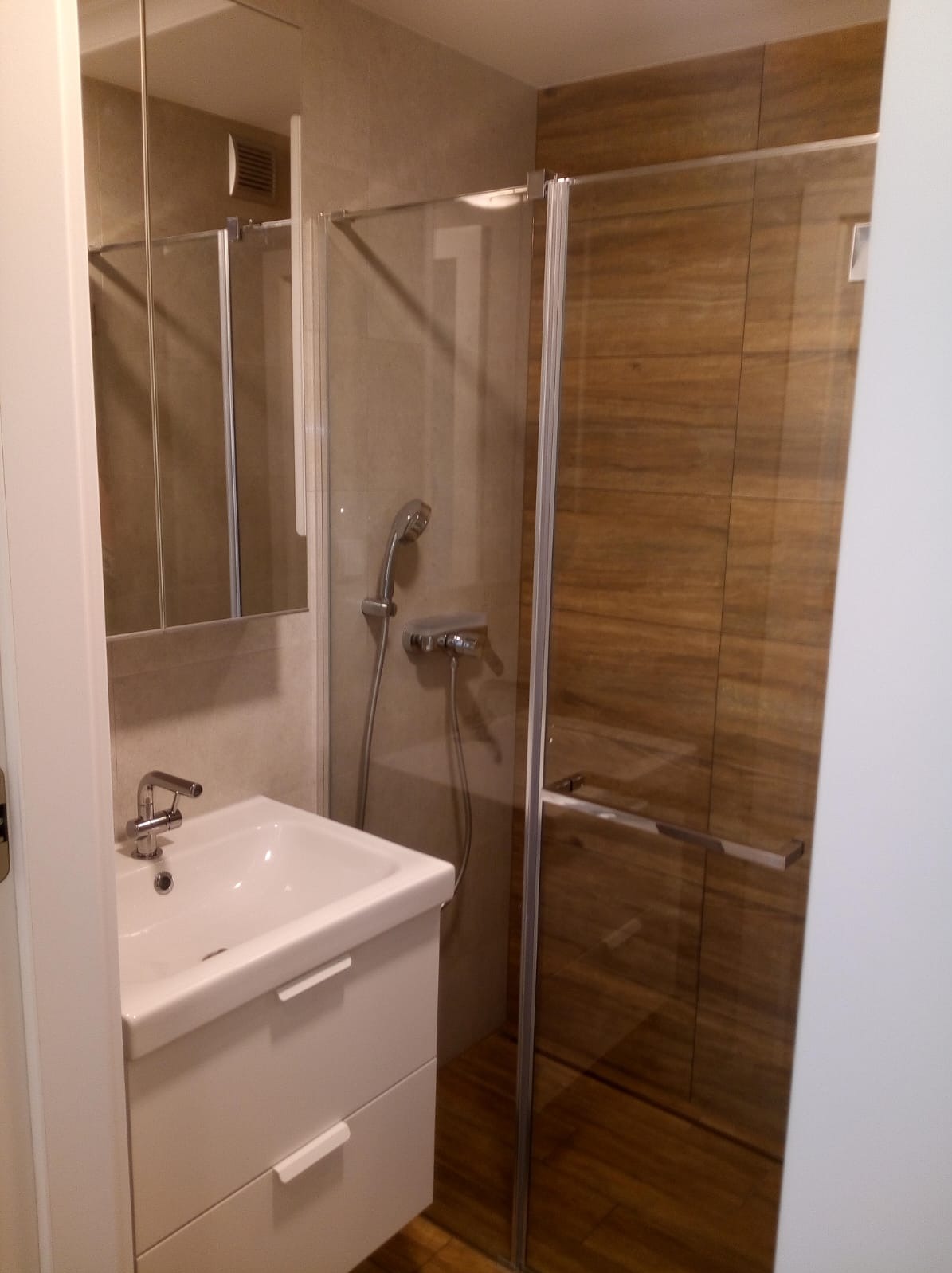 Nowoczesna łazienka z kabiną prysznicową typu walk-in, białą szafką pod umywalką i lustrem. Widoczne drewnopodobne płytki na ścianie kabiny.