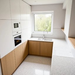 SZAJBA SPÓŁKA Z OGRANICZONĄ ODPOWIEDZIALNOŚCIĄ - Jasna kuchnia z białymi szafkami górnymi i drewnianymi dolnymi, okno z widokiem na zieleń, blat z białego lastryko. Minimalistyczny design, nowoczesne oświetlenie.