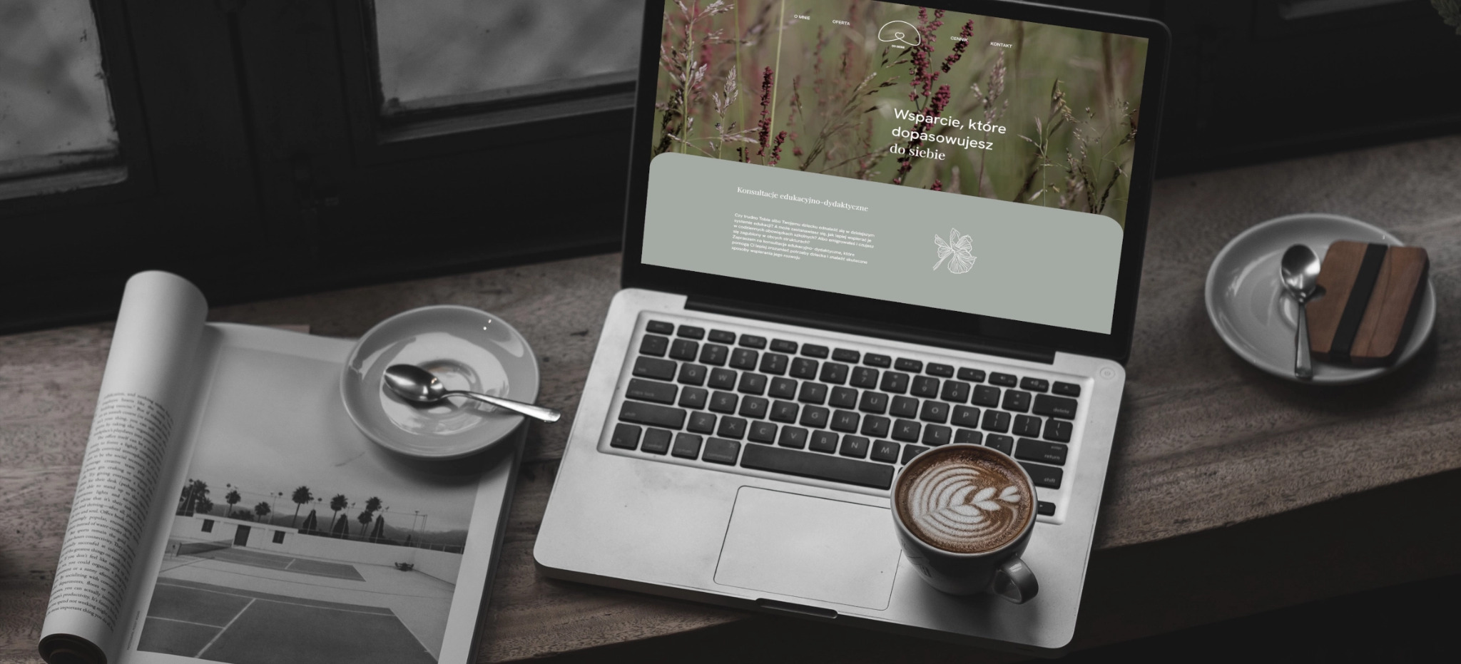 Laptop wyświetla stronę pomiedzyslowami.pl, filiżanka kawy i otwarty magazyn na drewnianym stole przy oknie. Prezentacja realizacji strony internetowej.