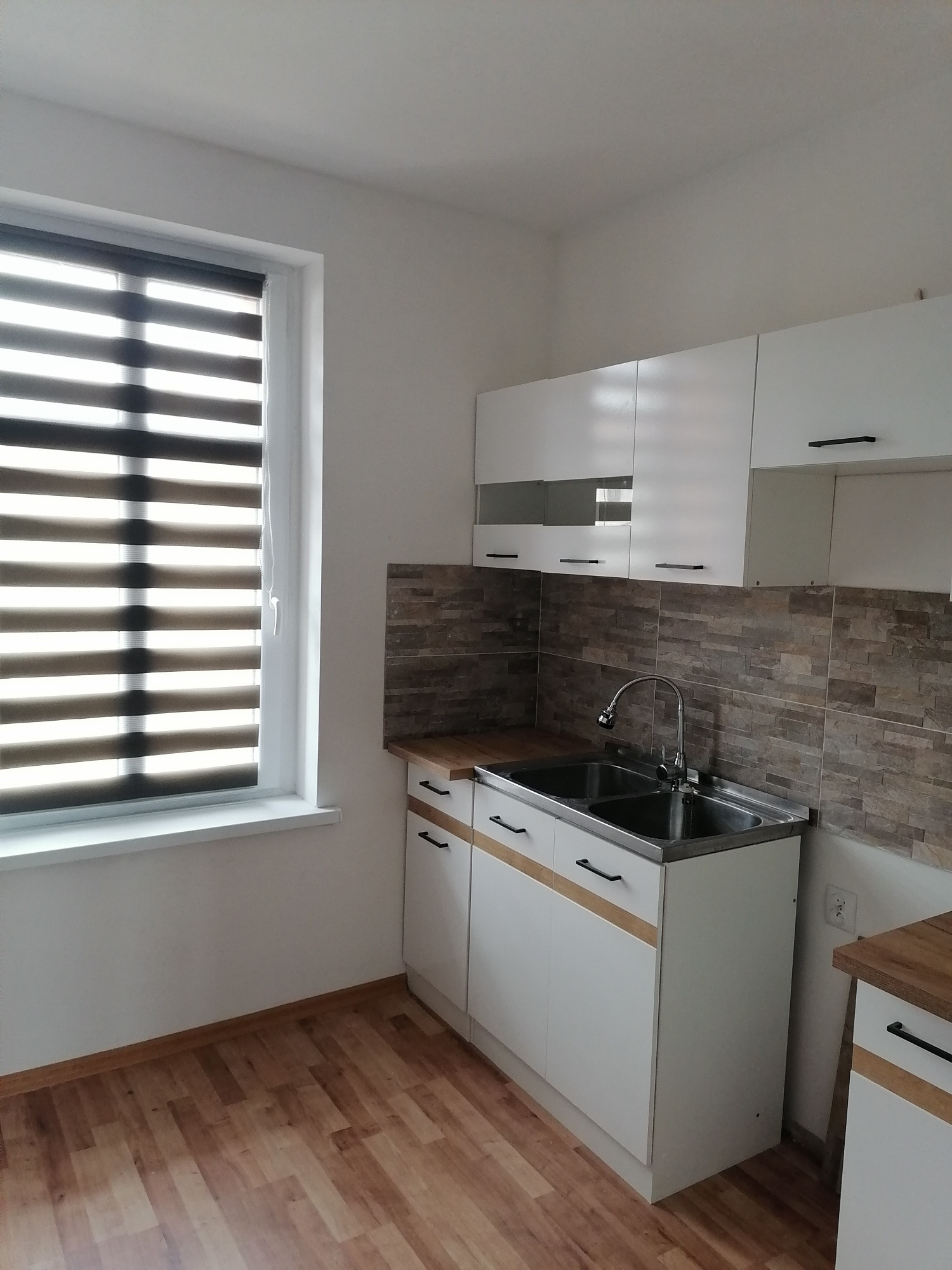 Jasna kuchnia z drewnianą podłogą, białymi szafkami z czarnymi uchwytami, stalowym zlewem i okładziną ścienną imitującą cegłę. Okno z żaluzjami.