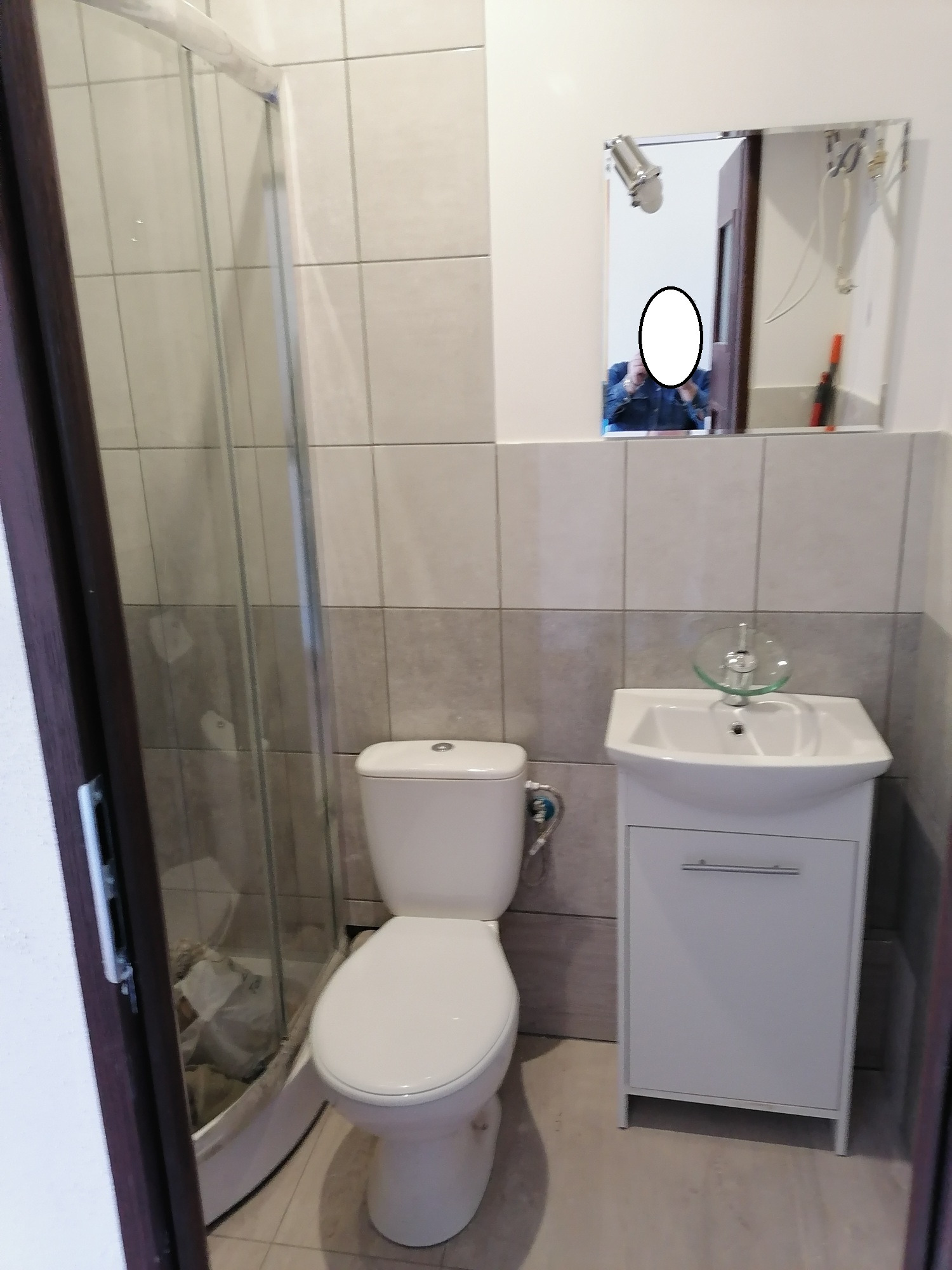 Wykończona łazienka z kabiną prysznicową, toaletą i umywalką z szafką. W lustrze odbija się osoba robiąca zdjęcie. Jasne płytki na ścianach.