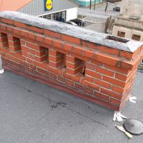 Ceglana attyka z otworami na dachu budynku, widok z góry. Widoczne fragmenty dachu pokrytego papą i budynki w tle. Nietypowe wykończenie detalu architektonicznego.