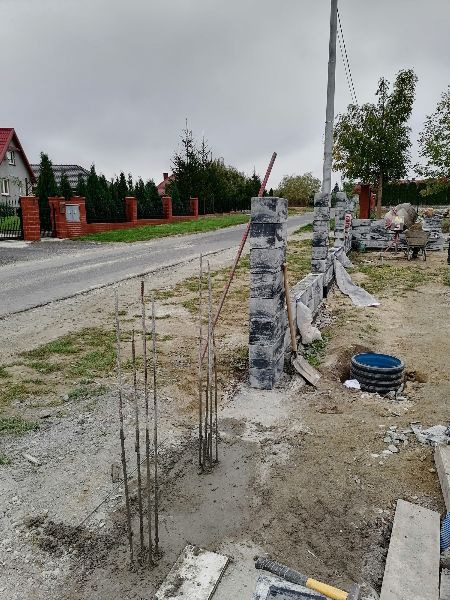 Budowa ogrodzenia z bloczków betonowych: widoczne fundamenty z wystającą stalą zbrojeniową, częściowo wzniesione słupki i fragment muru. W tle dom i ulica.